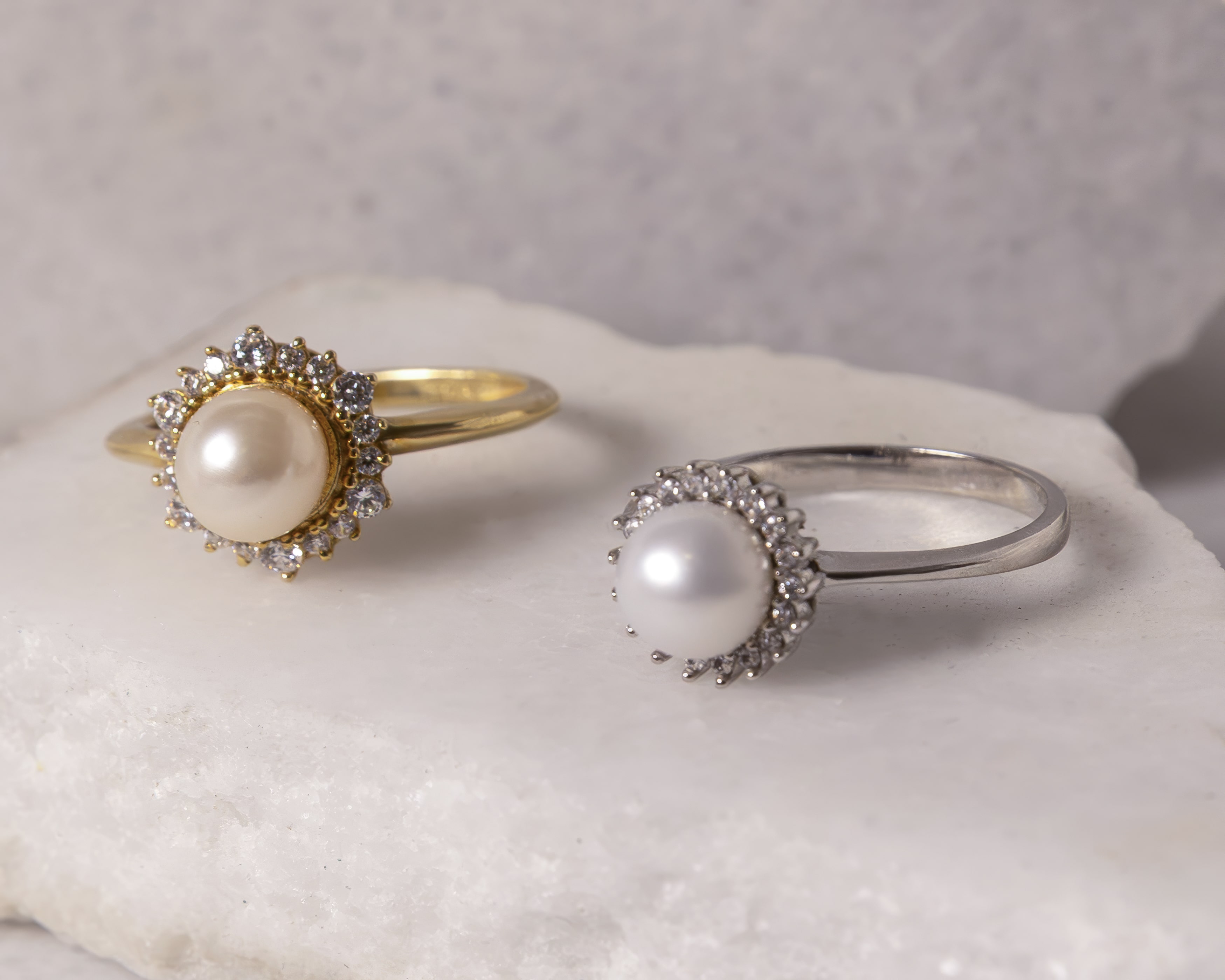Two pearl rings on a white stone surface, one gold with a cream pearl and one silver with a white pearl, both surrounded by small diamonds.