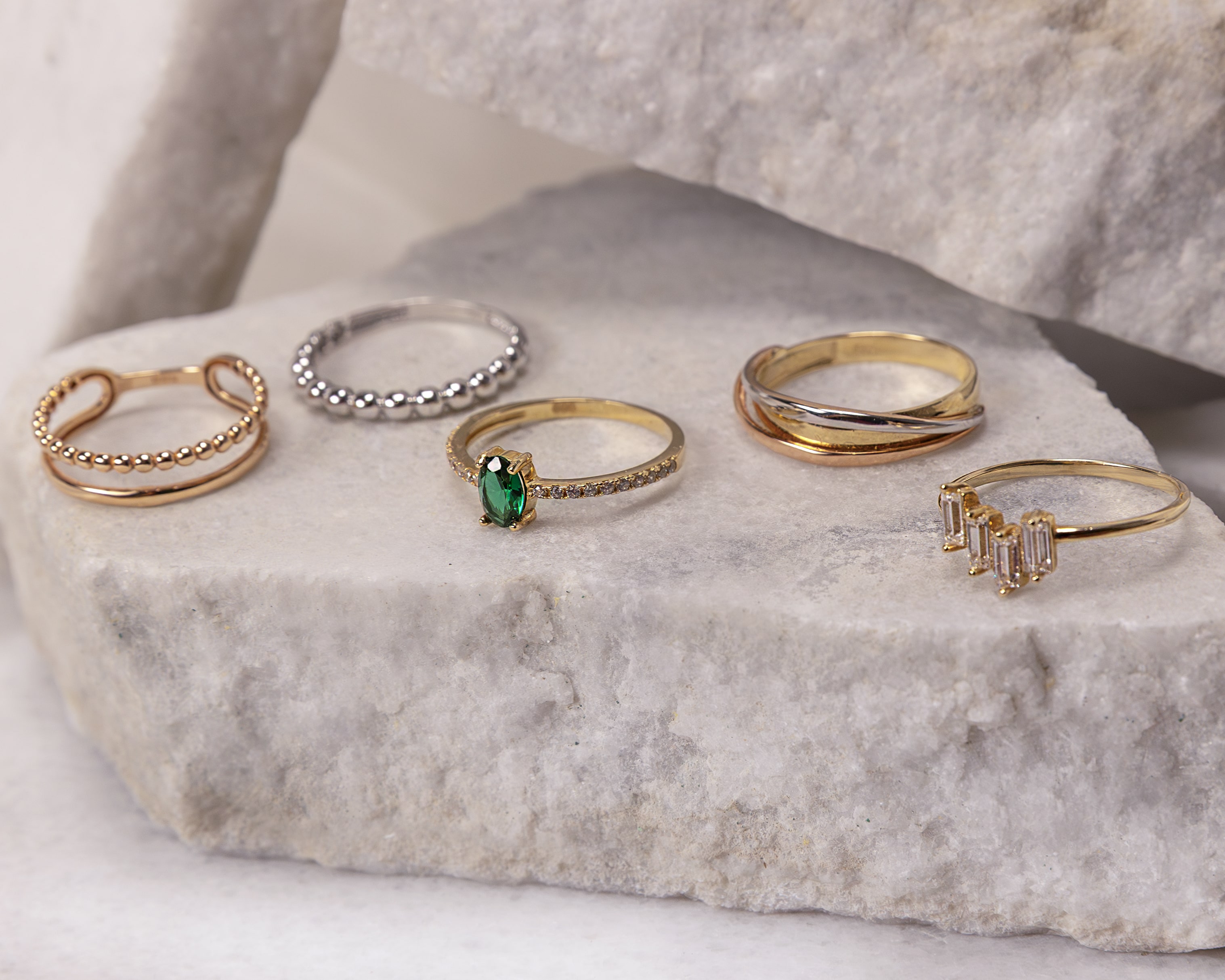 Five elegant rings displayed on a rough stone surface, featuring gold and silver tones with one ring adorned with a green gemstone.