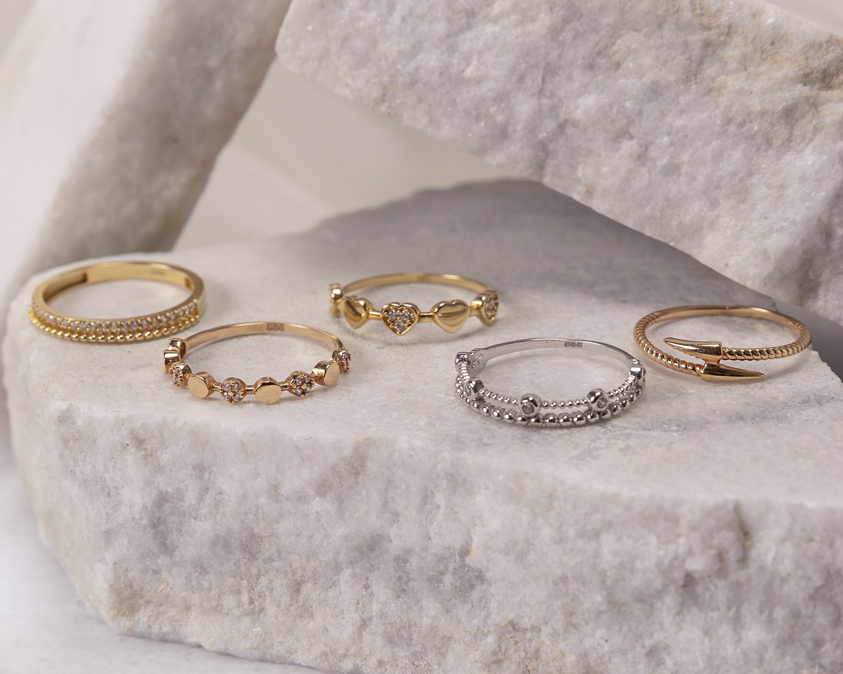Five delicate rings with gold and silver tones displayed on textured stone blocks, featuring designs with small gems, hearts, and pointed tips.