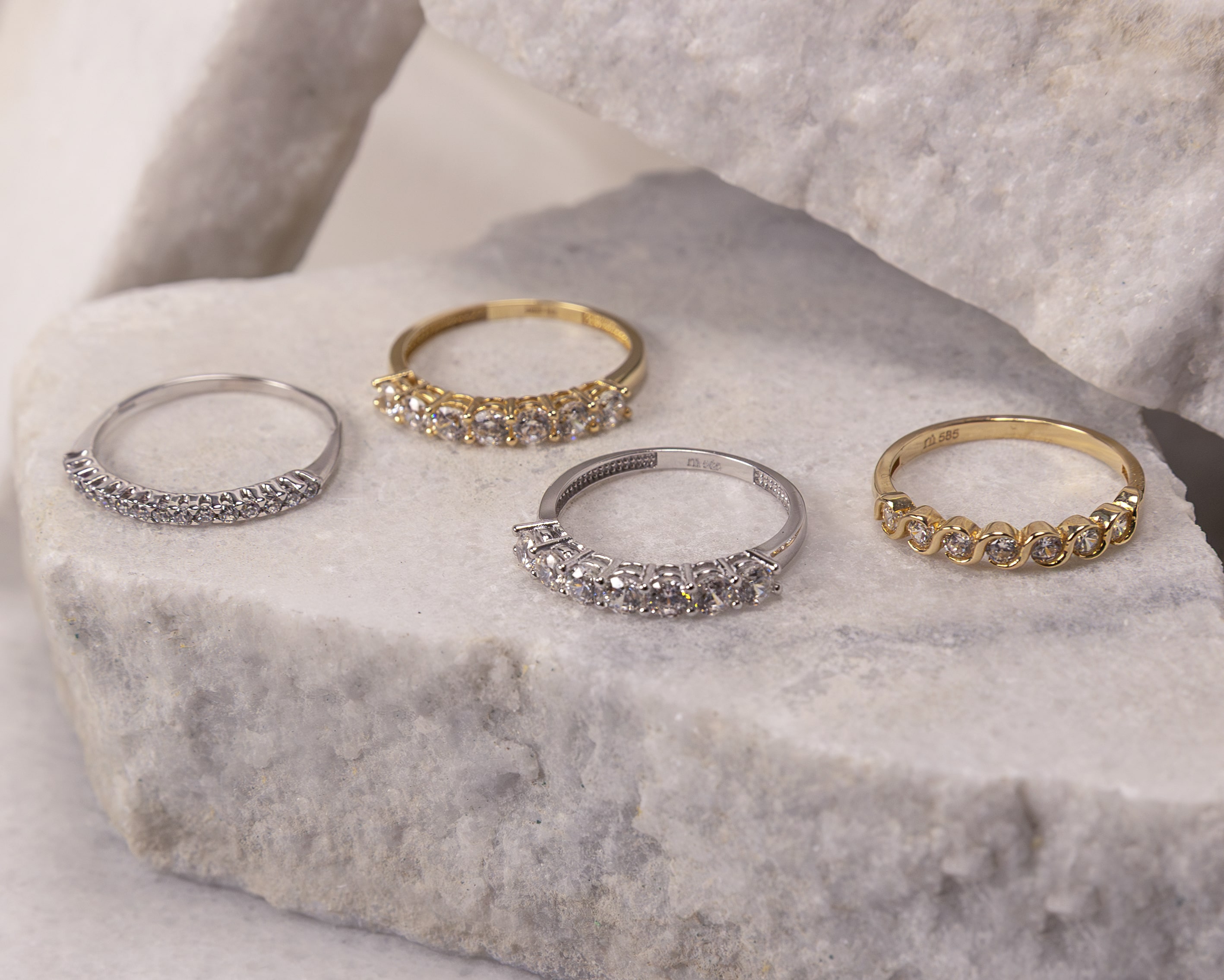 Four gold and silver rings with rows of small diamonds displayed on textured beige stone slabs.