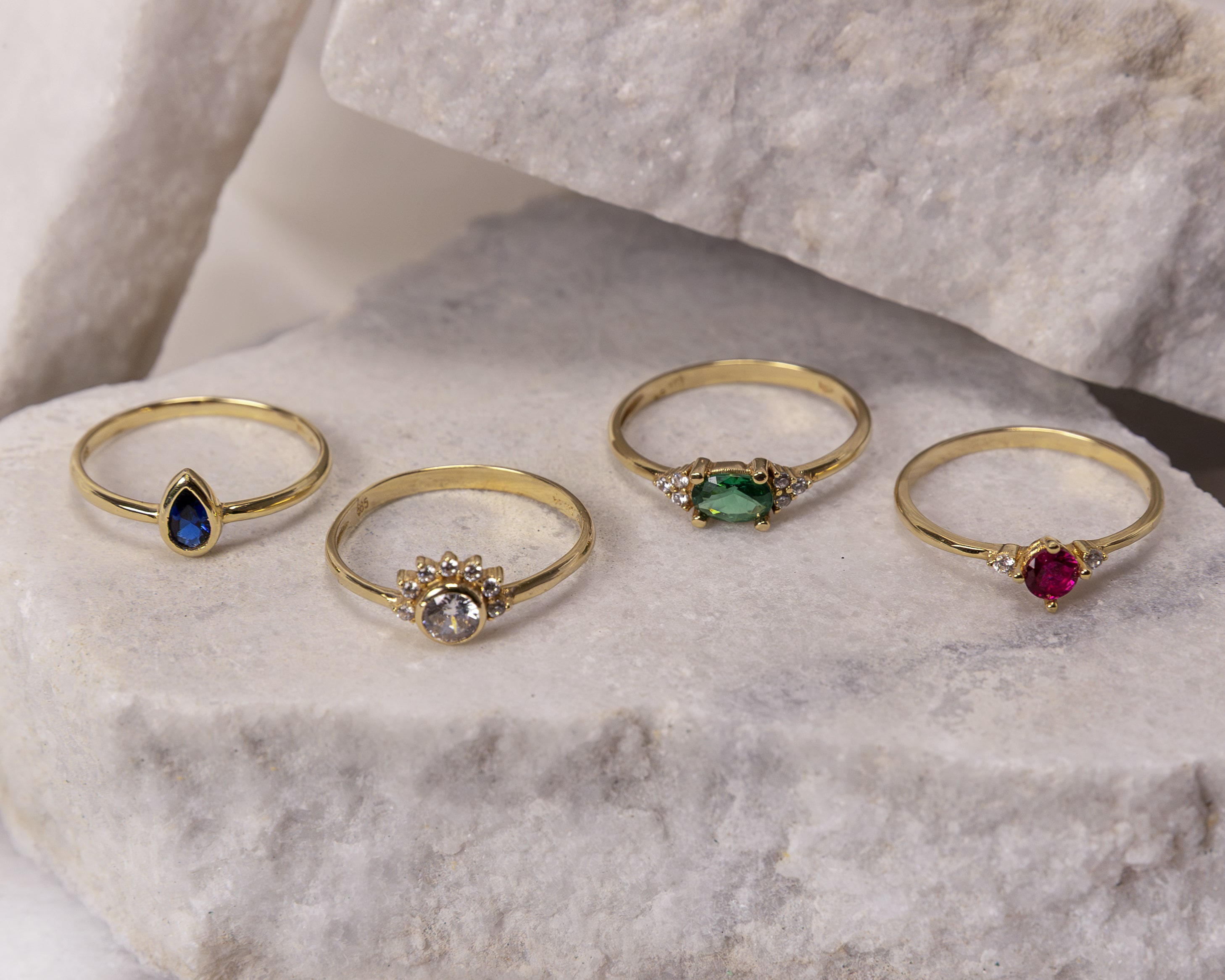 Four gold rings with different gemstones—blue pear-shaped, clear round with small diamonds, green oval, and red round—on light gray stone slabs.
