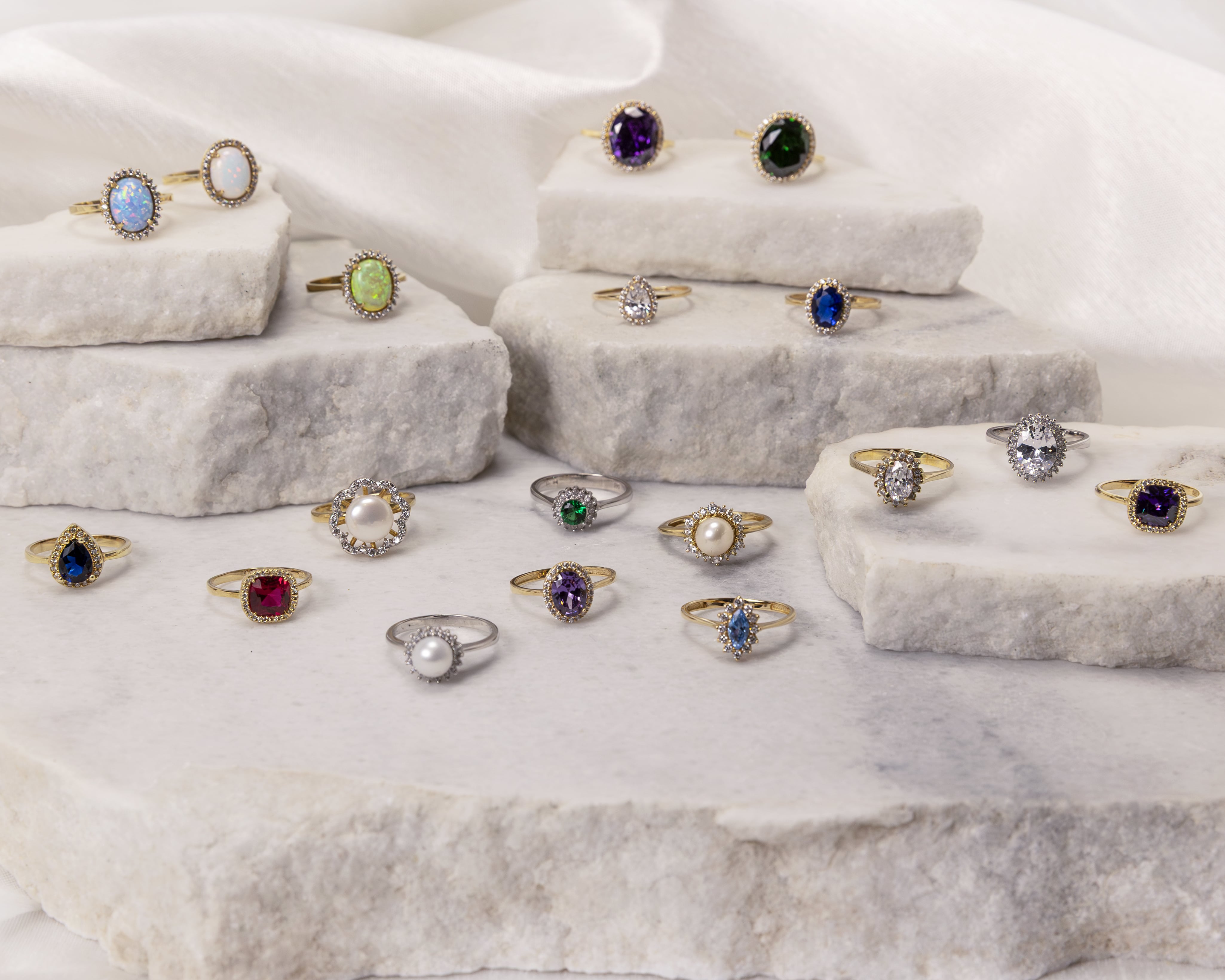 Assortment of gold and silver rings with colorful gemstones and pearls displayed on stacked white stone slabs.
