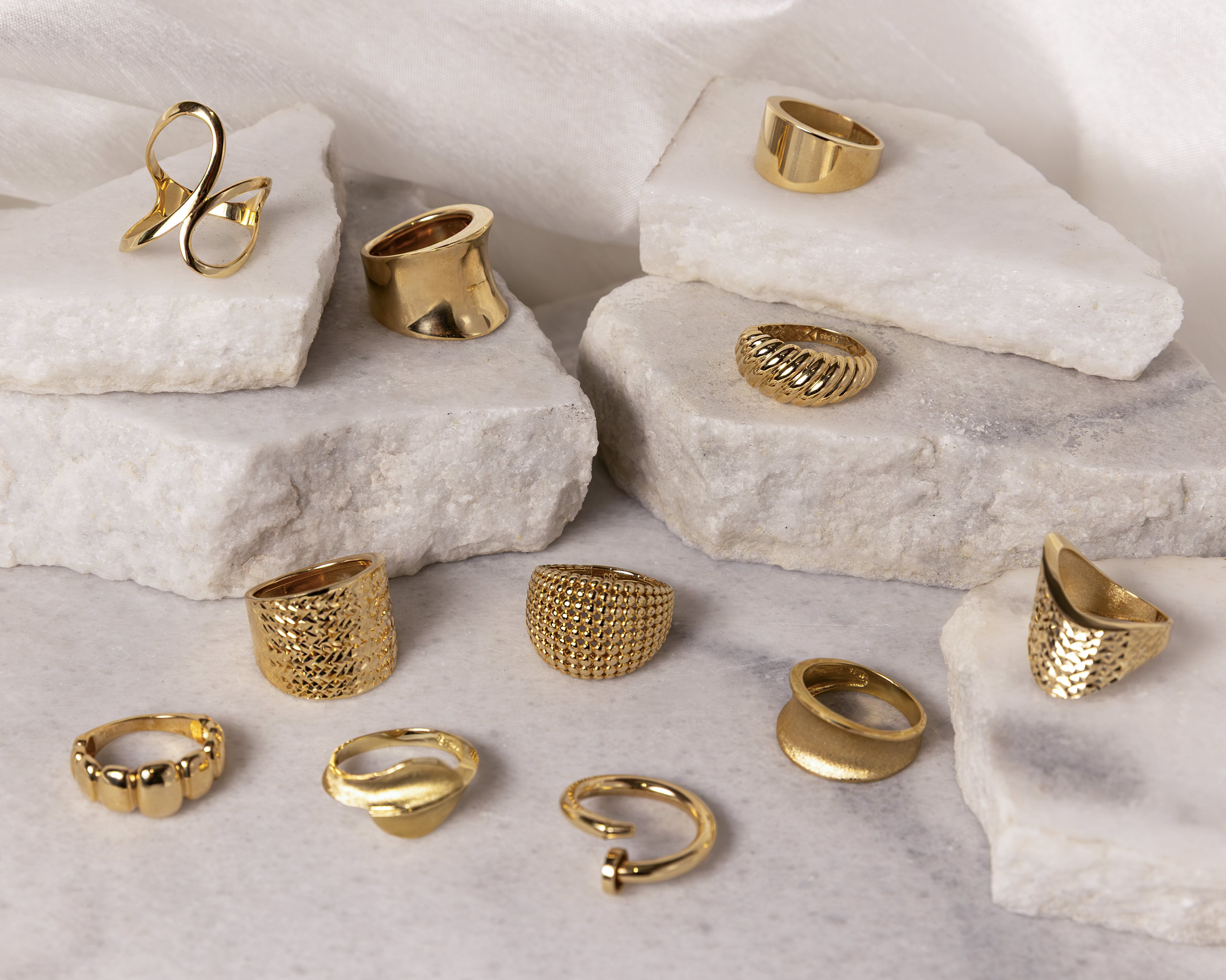Twelve assorted gold rings with various textures and designs displayed on layered white marble slabs.