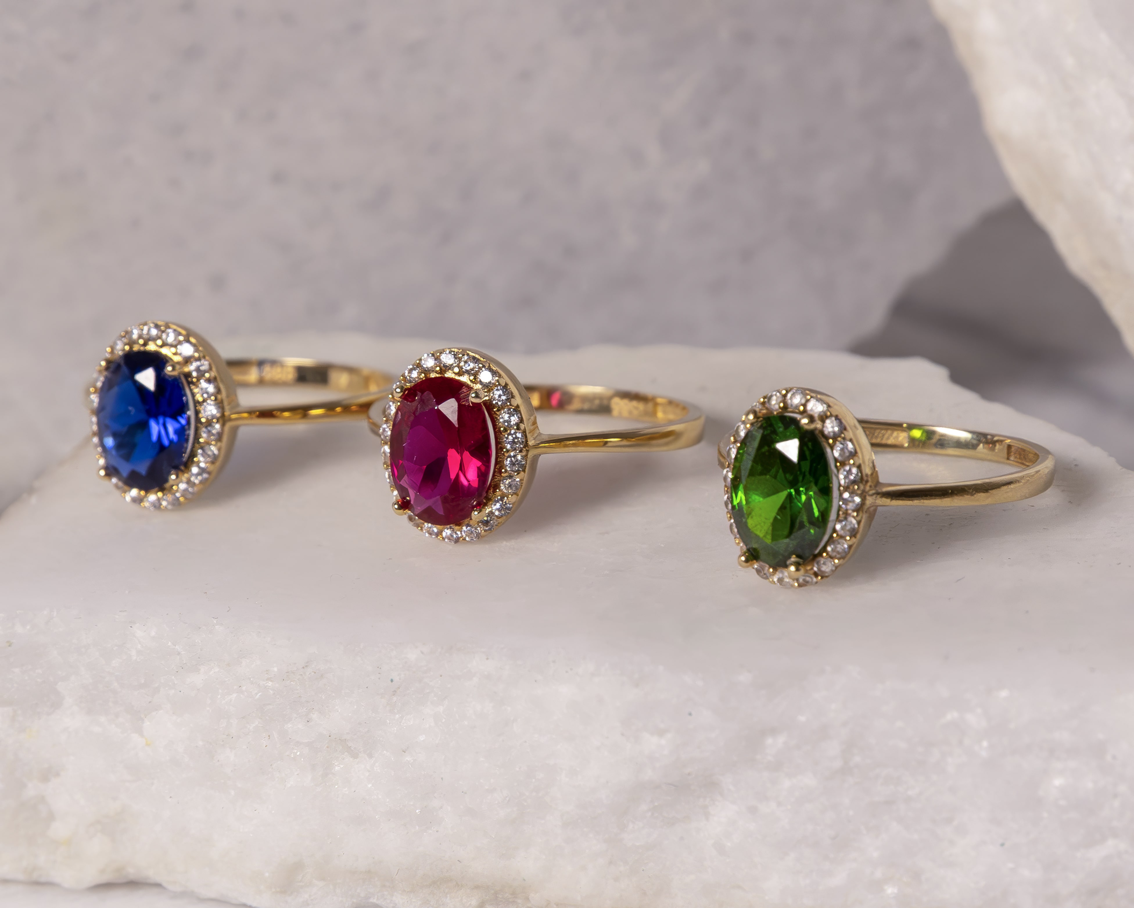 Three gold rings with oval gemstones in blue, red, and green, each surrounded by small white diamonds, displayed on a light stone surface.
