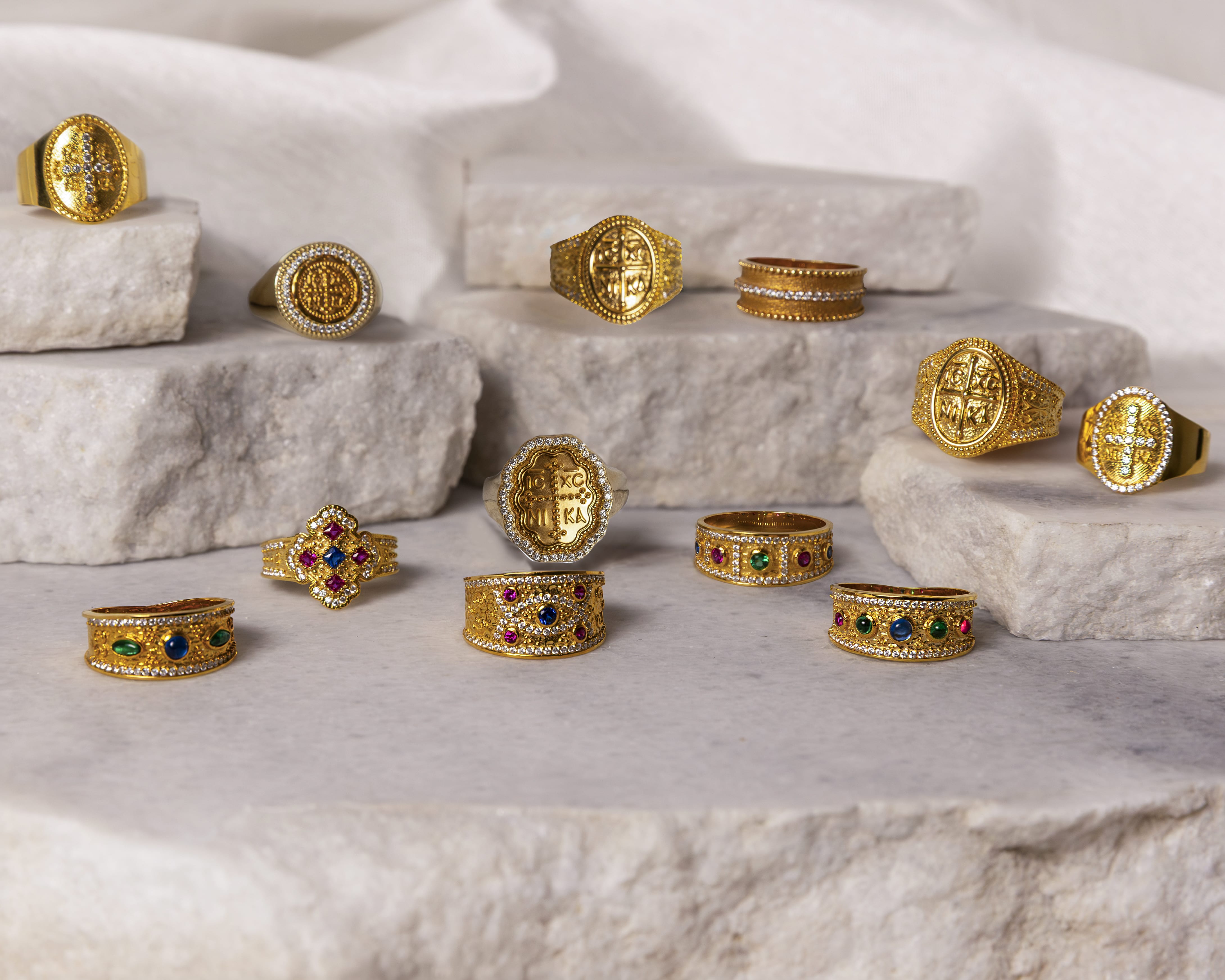Collection of ornate gold rings with colorful gemstones and intricate designs displayed on white stone slabs.