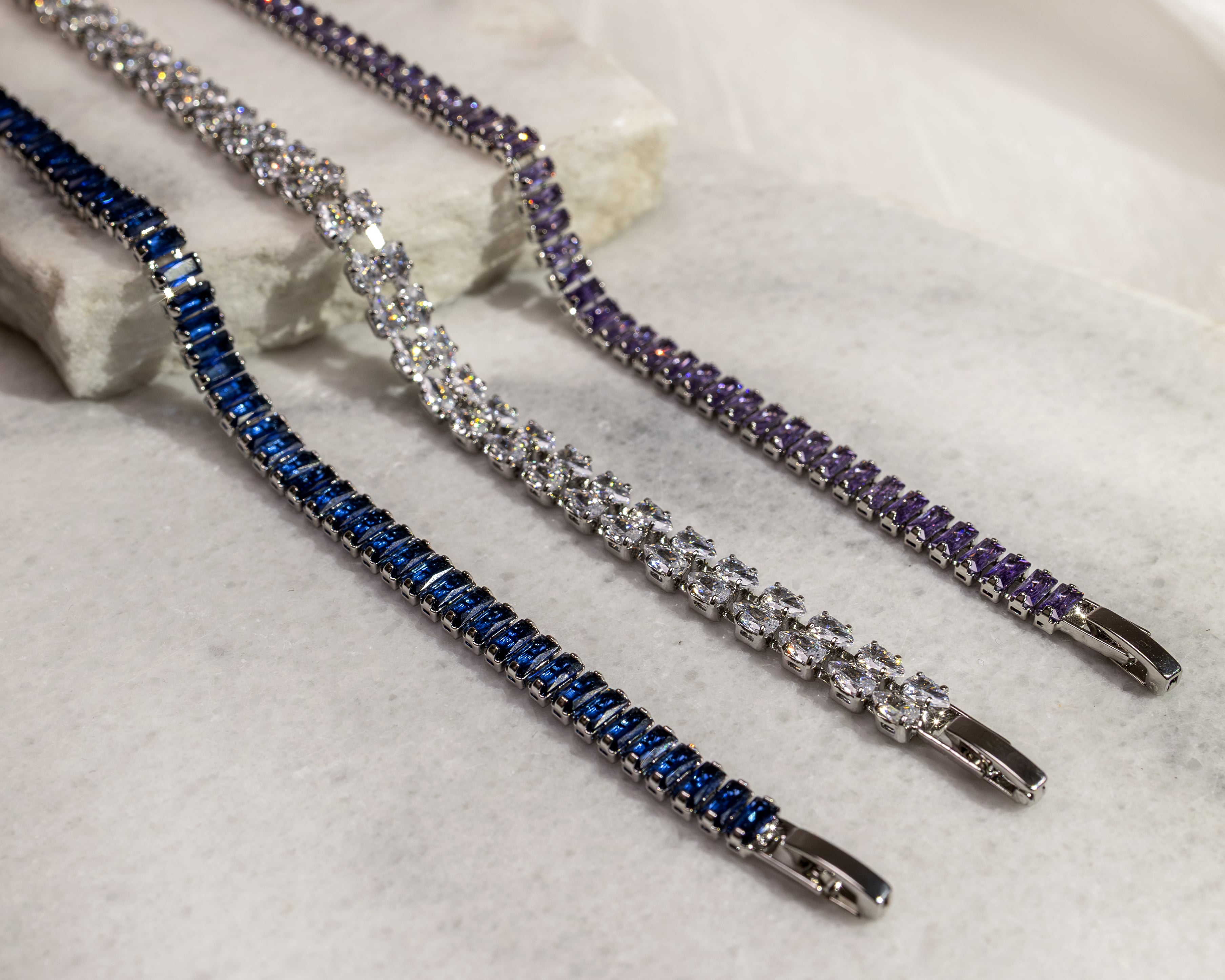 Three silver bracelets with blue, clear, and purple gemstones arranged on a light marble surface.