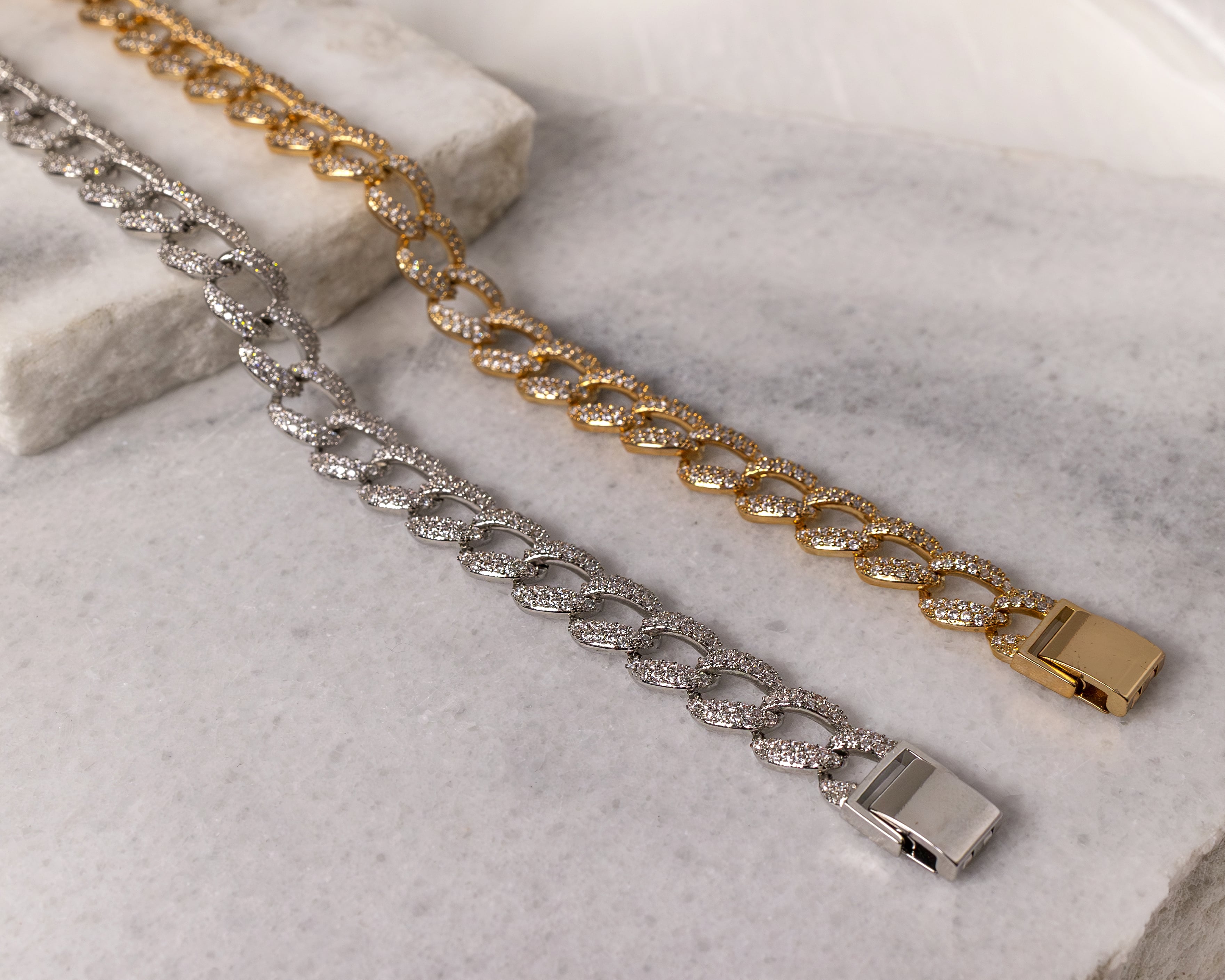 Two chain bracelets with sparkling stones, one gold and one silver, displayed on a marble surface.