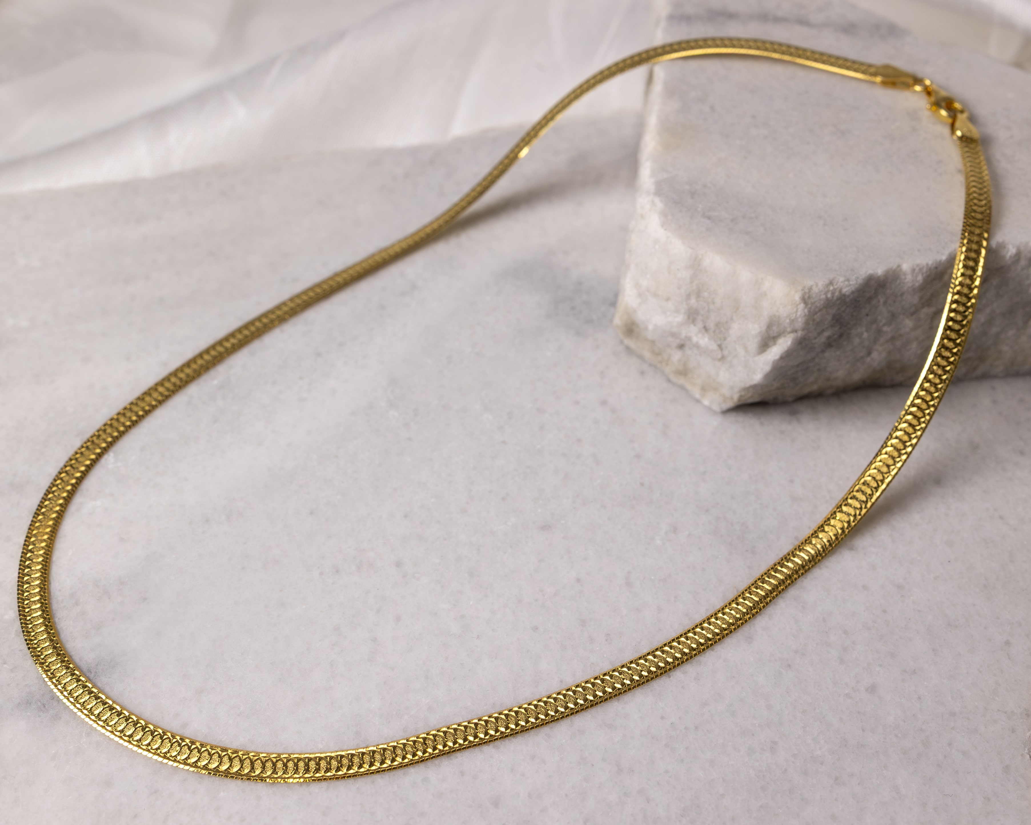 Gold chain necklace with a textured pattern laid out on a light marble surface near a rough stone block.