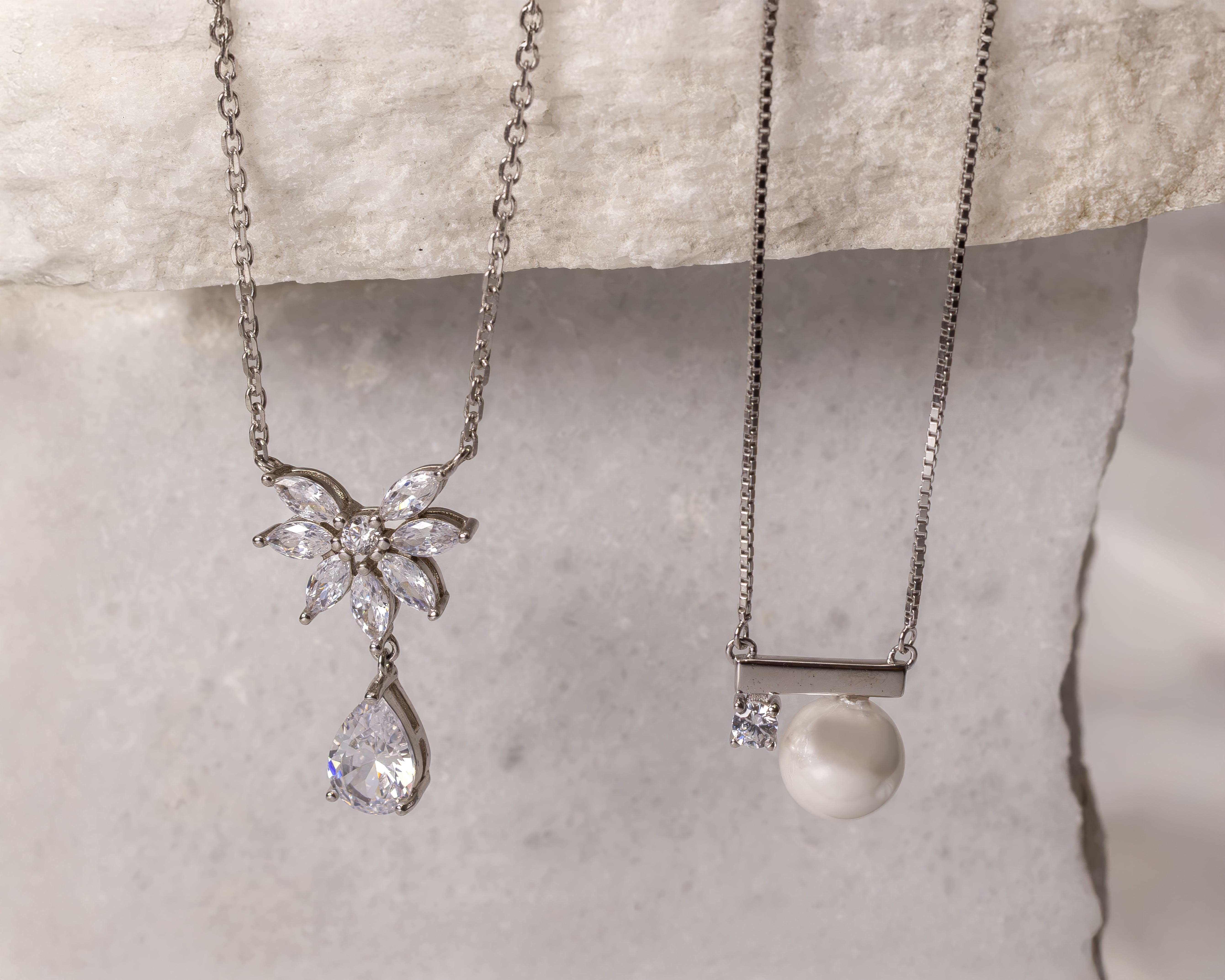 Two silver necklaces hanging against a textured stone background, one with a flower-shaped crystal pendant and a teardrop crystal, the other with a pearl and small square crystal pendant.