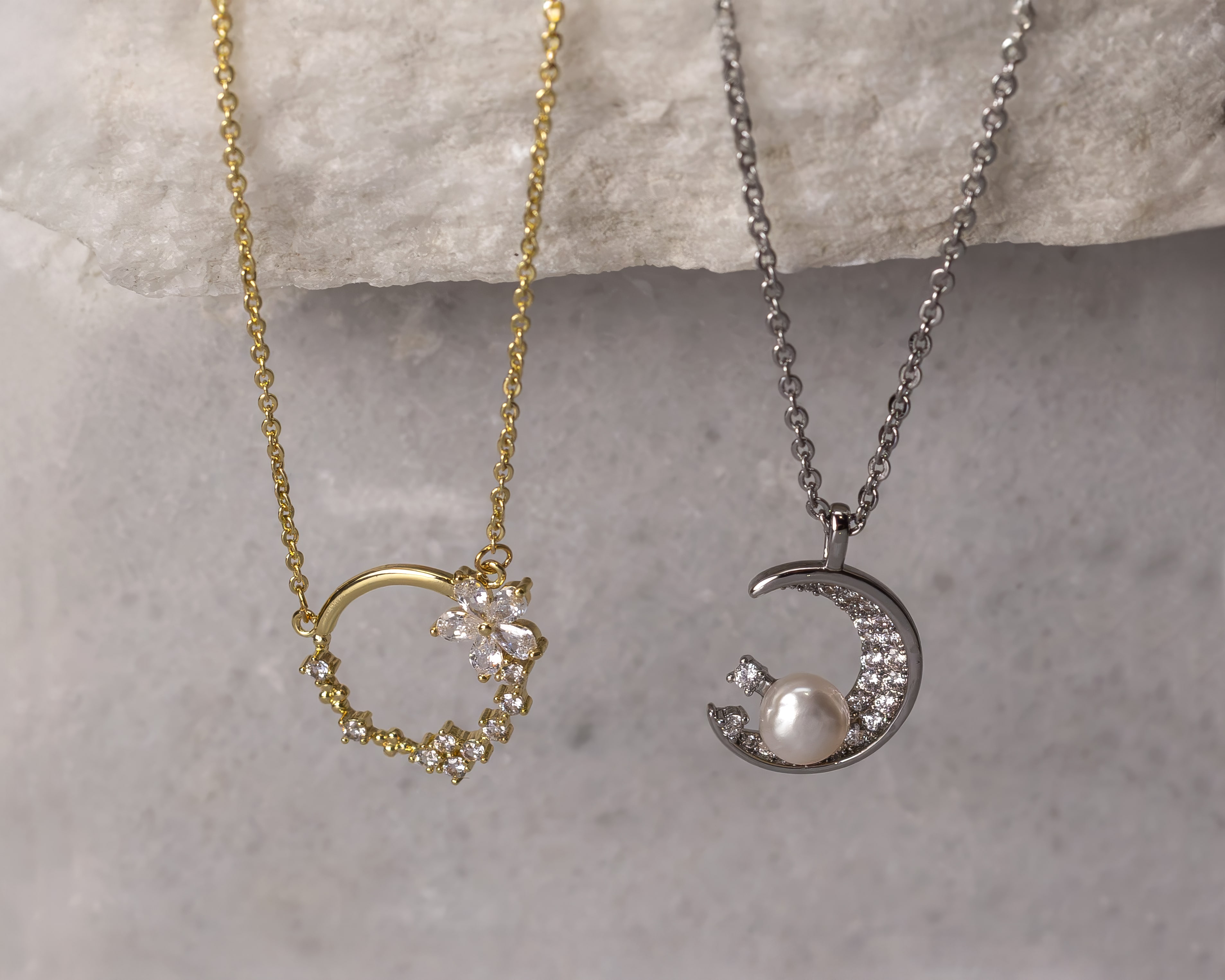 Two delicate necklaces on a stone surface; one gold heart-shaped pendant with small crystals and a flower, the other a silver crescent moon pendant with embedded crystals and a pearl.