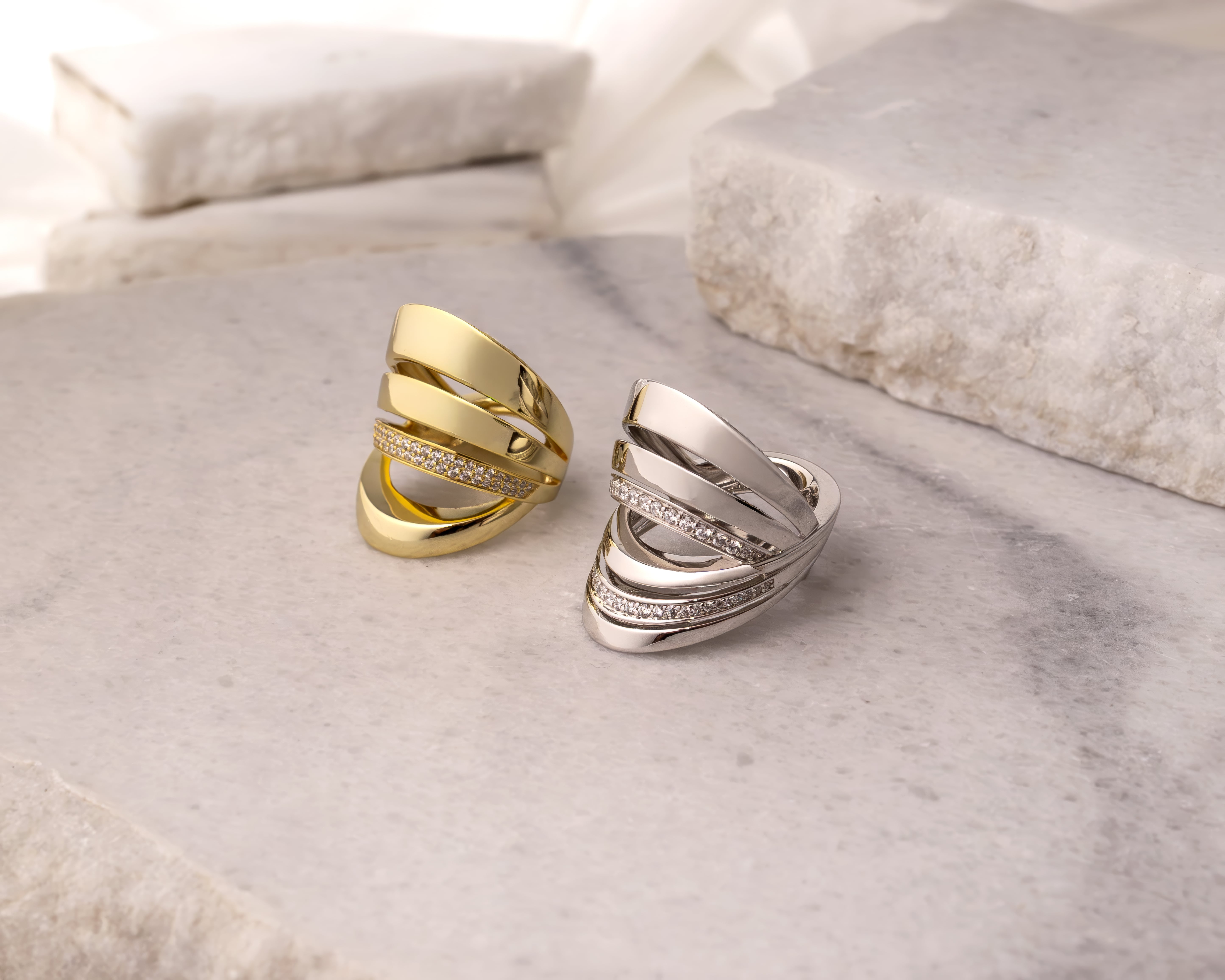 Two wide, multi-band rings with embedded small diamonds, one gold and one silver, displayed on a marble surface.