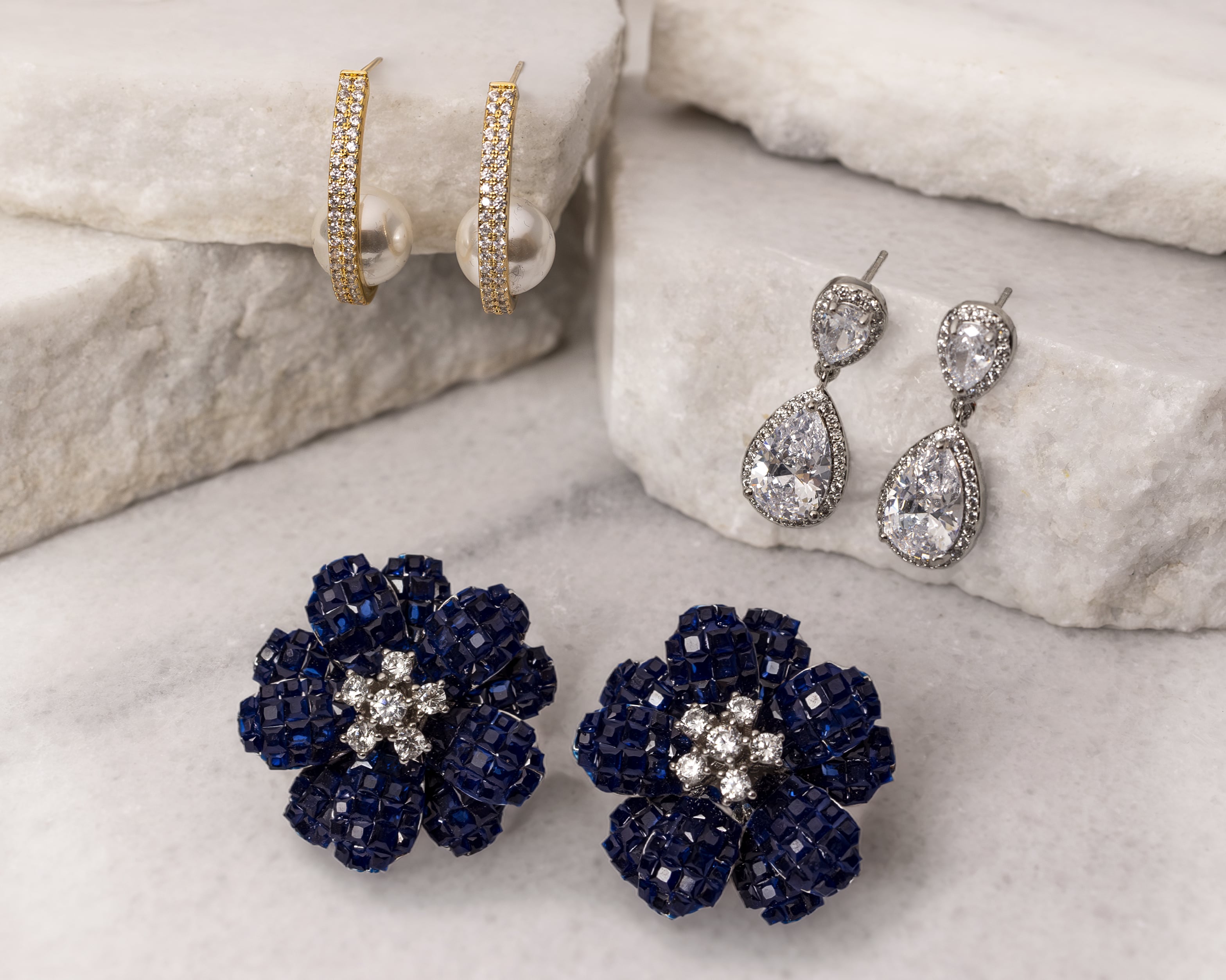 Three pairs of elegant earrings on white stone: gold hoops with pearls and diamonds, silver teardrop diamond studs, and blue jeweled flower studs with diamond centers.