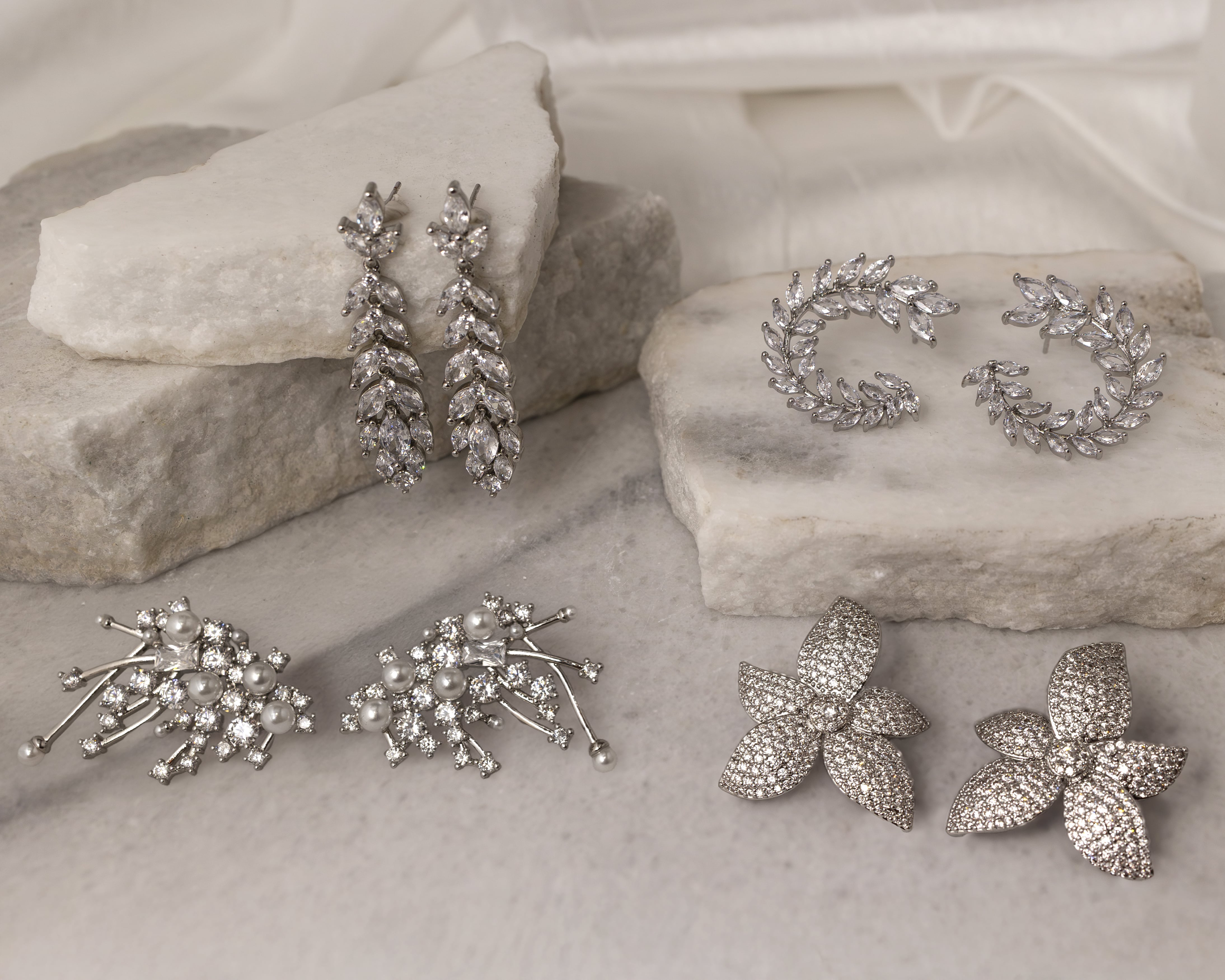 Four pairs of silver and crystal earrings displayed on white marble slabs, including leaf designs, floral clusters with pearls, and pavé flower shapes.