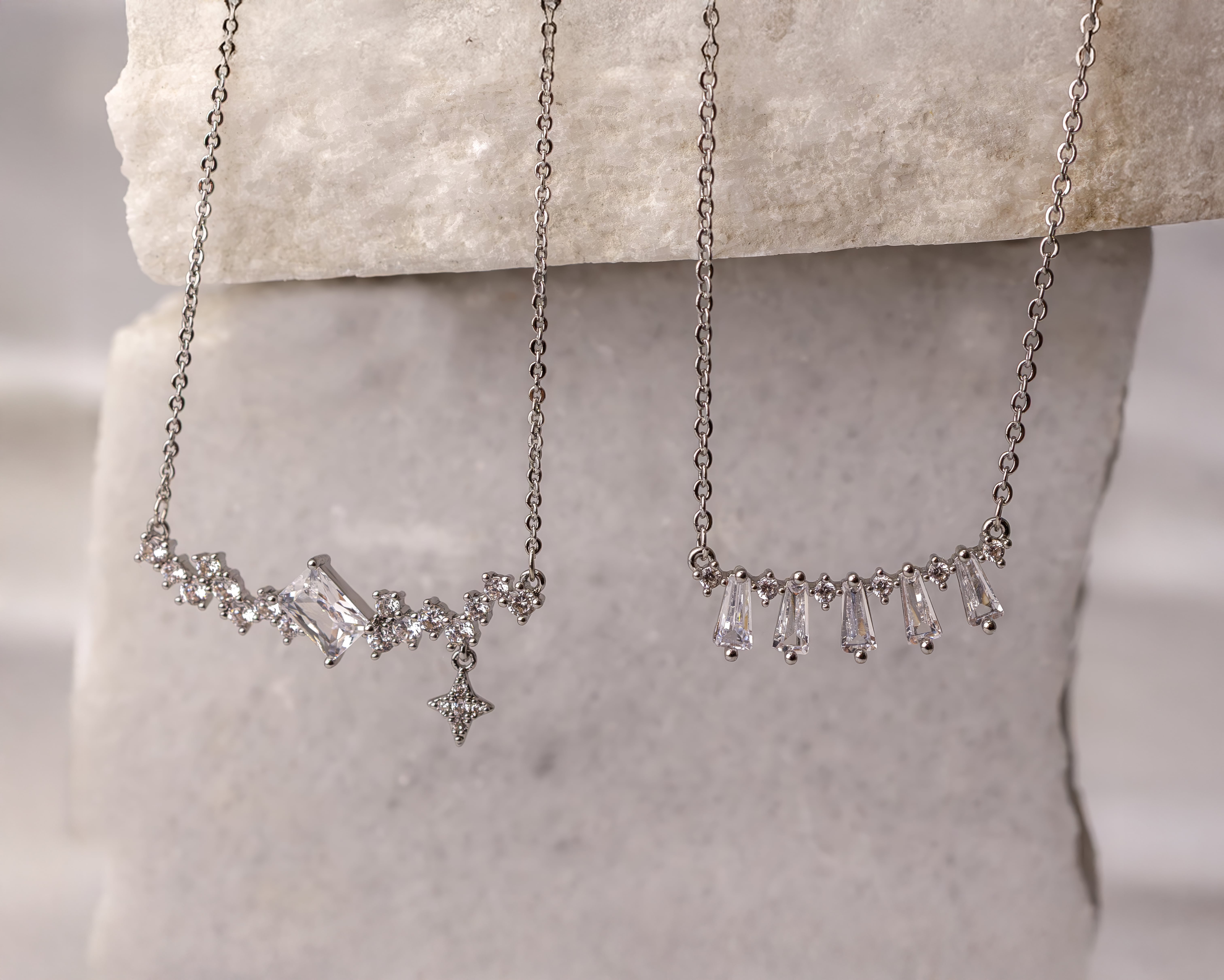 Two delicate silver necklaces with diamond-like gemstones displayed against a textured stone background.