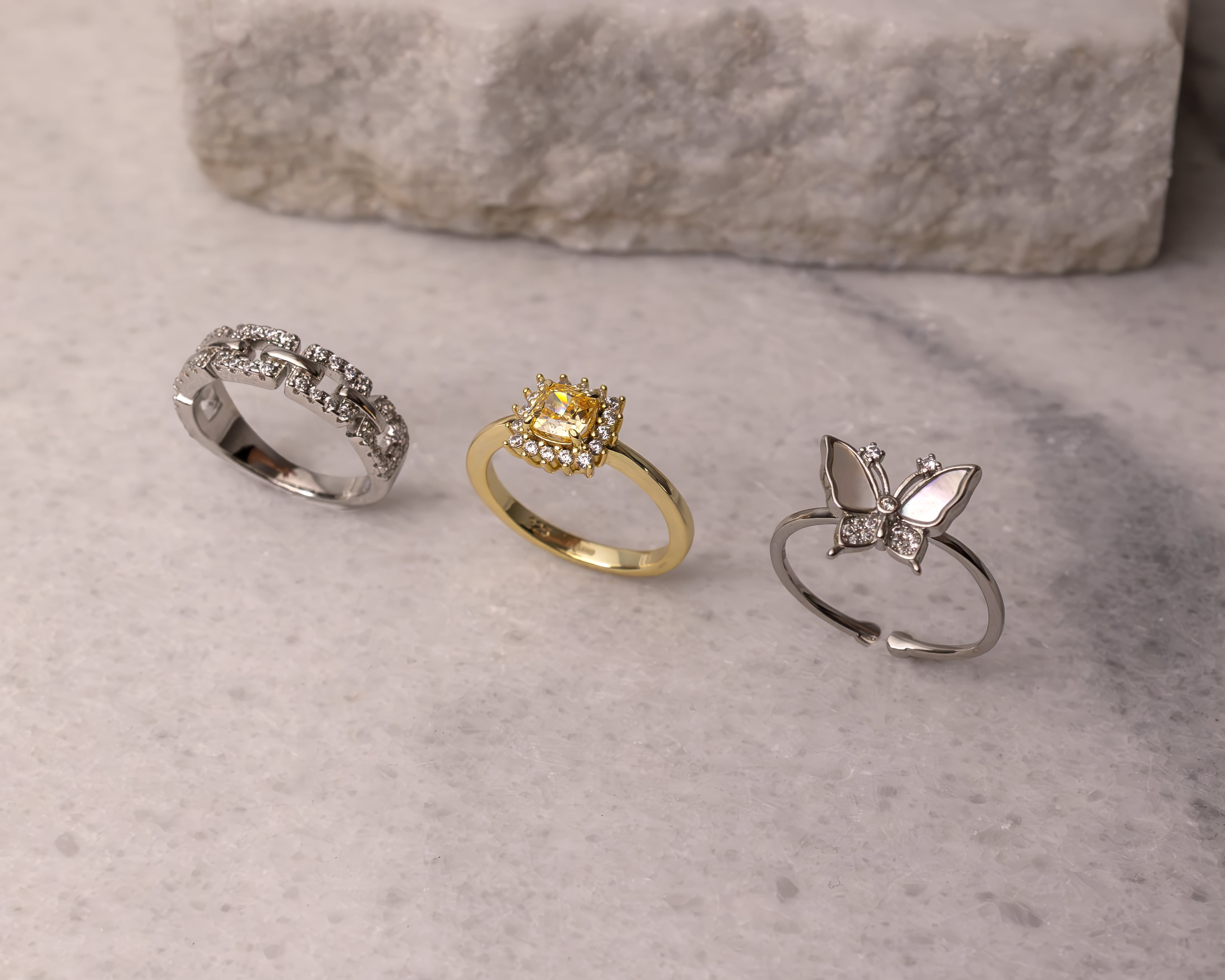 Three rings on a marble surface: a silver chain-link diamond ring, a gold ring with a yellow gemstone surrounded by small diamonds, and a silver butterfly ring with pearl-like wings and diamond accents.