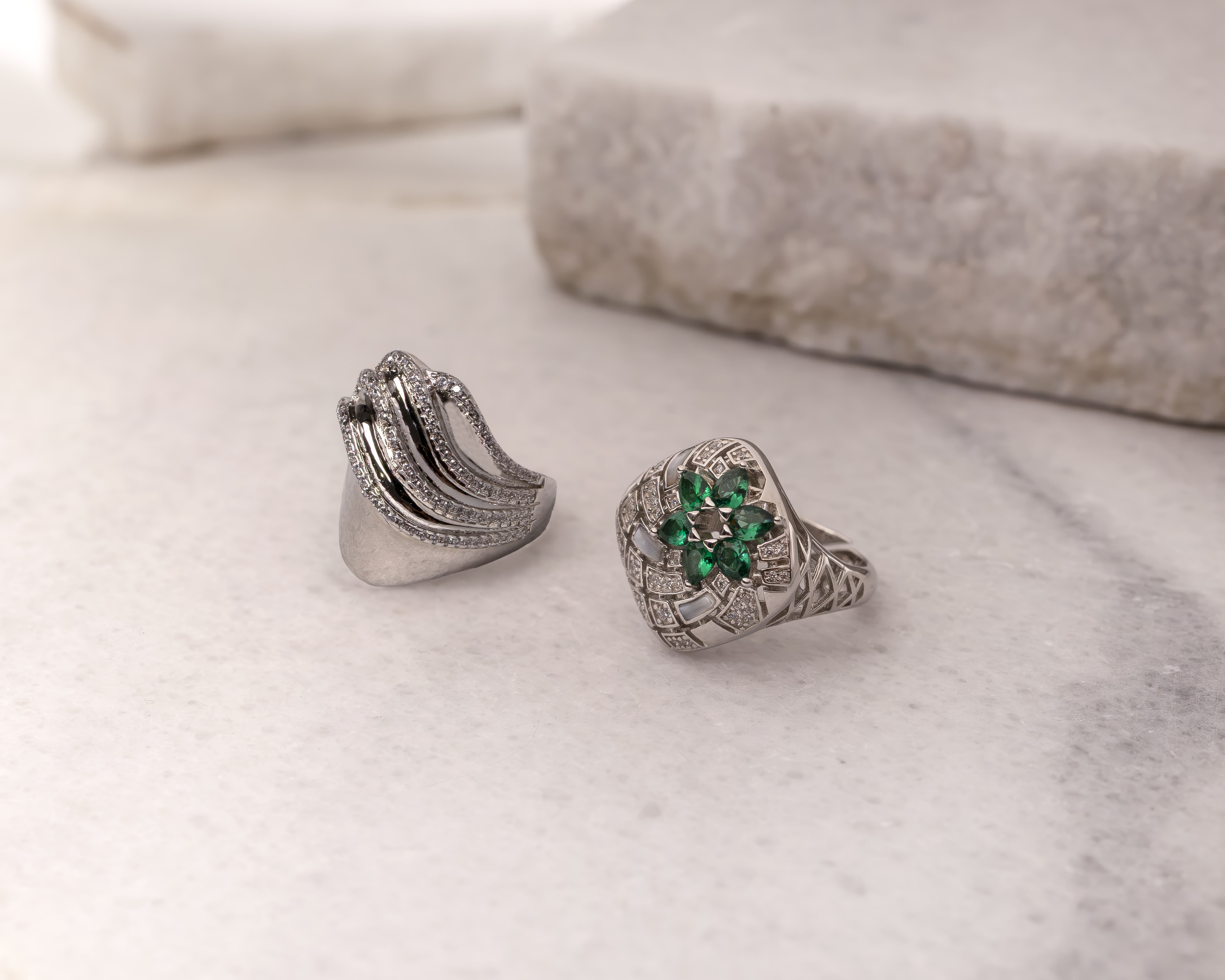Two ornate silver rings on a marble surface, one with a floral design of green gemstones and diamonds, the other with curved rows of small diamonds.