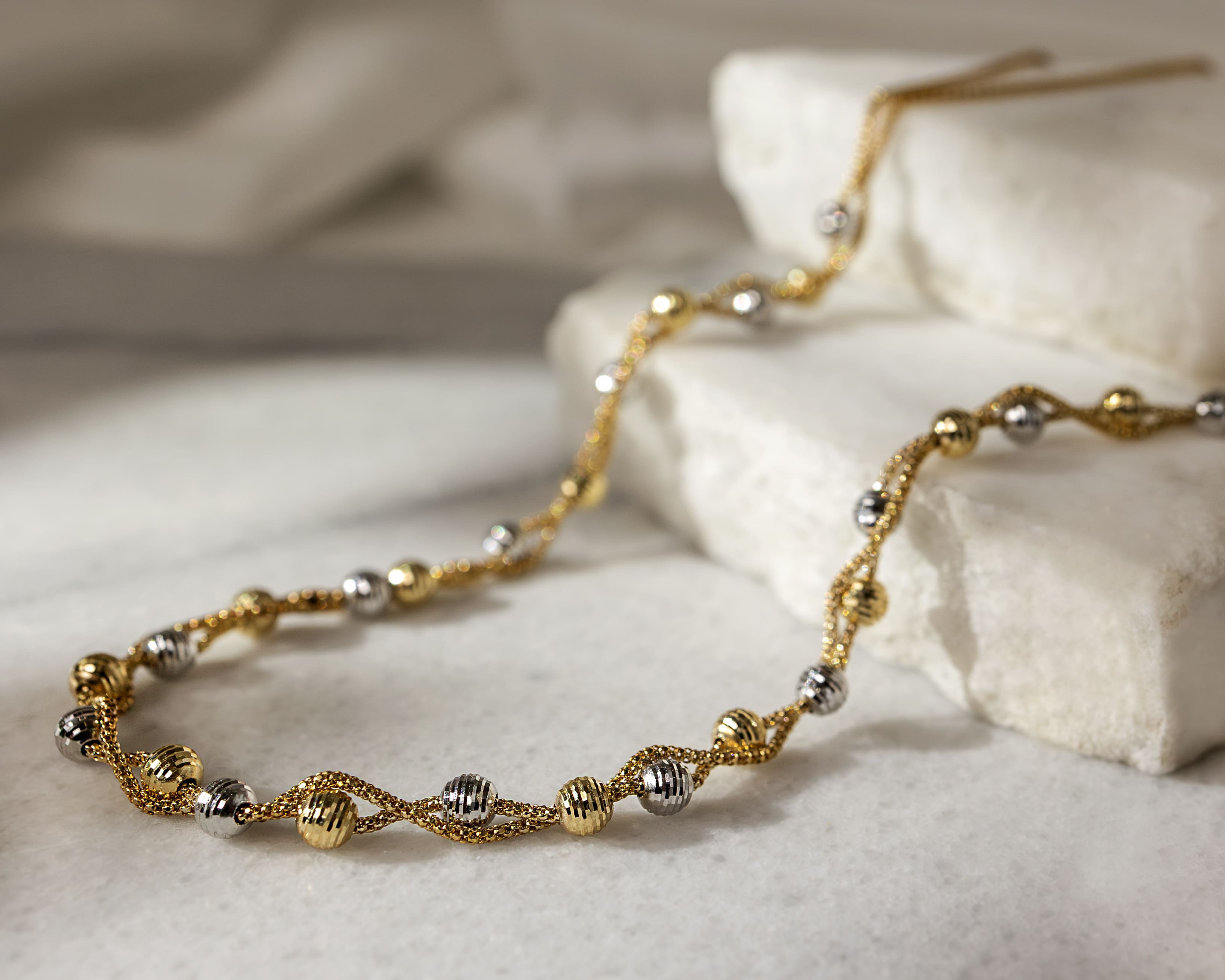 Close-up of a gold and silver twisted chain necklace with textured spherical beads on a marble surface.