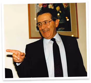 Uomo sorridente con occhiali, vestito con giacca scura, camicia bianca e cravatta nera che indica con la mano destra.