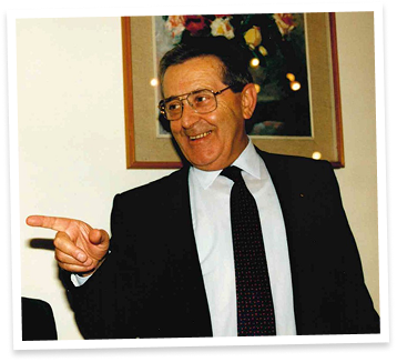 Uomo sorridente con occhiali, vestito con giacca scura, camicia bianca e cravatta nera che indica con la mano destra.