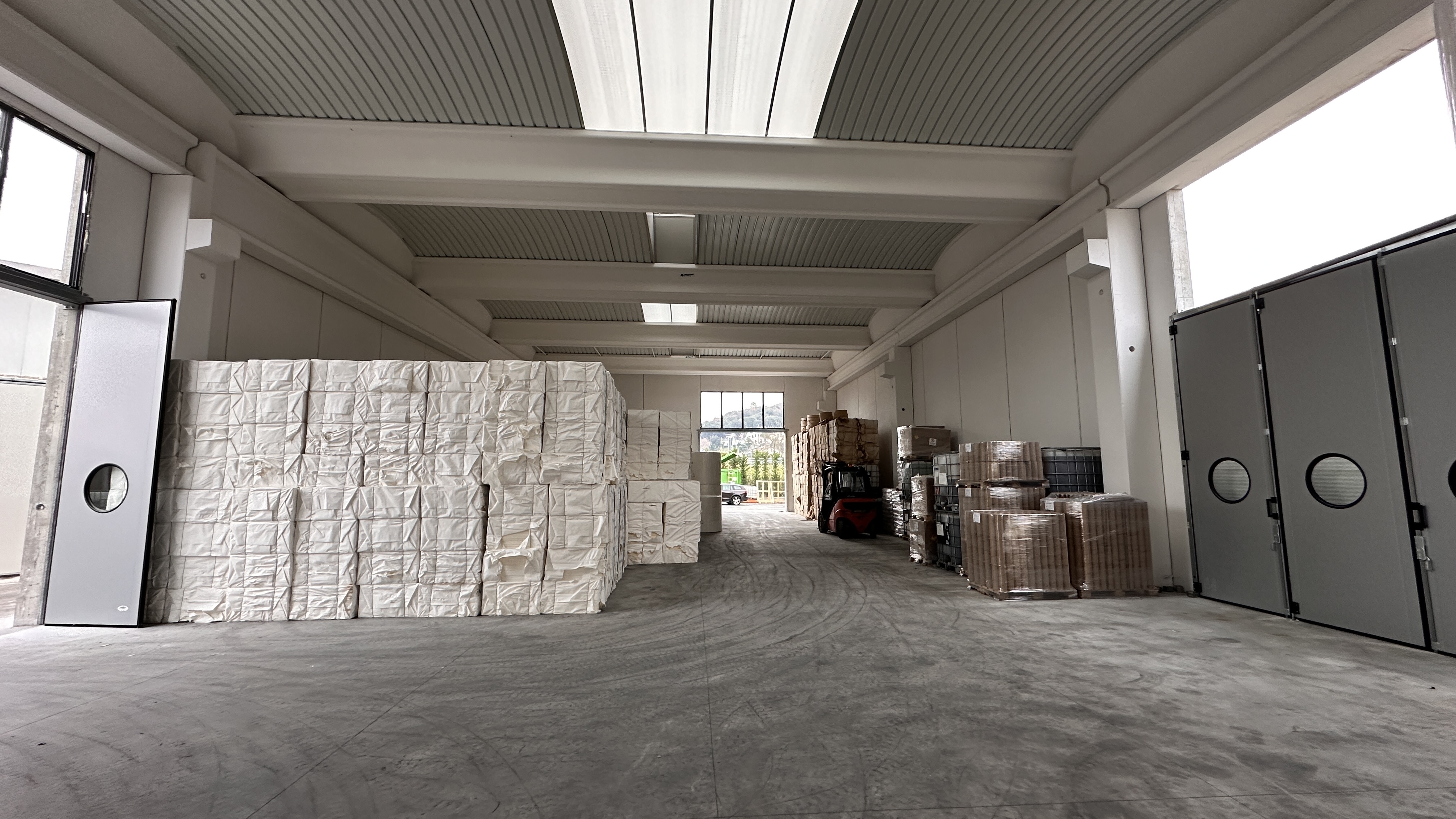 Magazzino con grandi pallet impilati coperti da teli bianchi a sinistra e pallet con scatole marroni e contenitori a destra, con un carrello elevatore al centro.