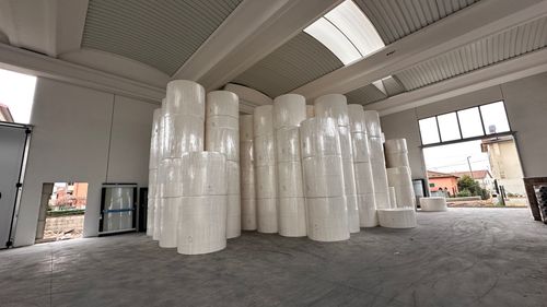 Grande magazzino con numerosi grandi rotoli di carta bianca impilati al centro su pavimento in cemento.