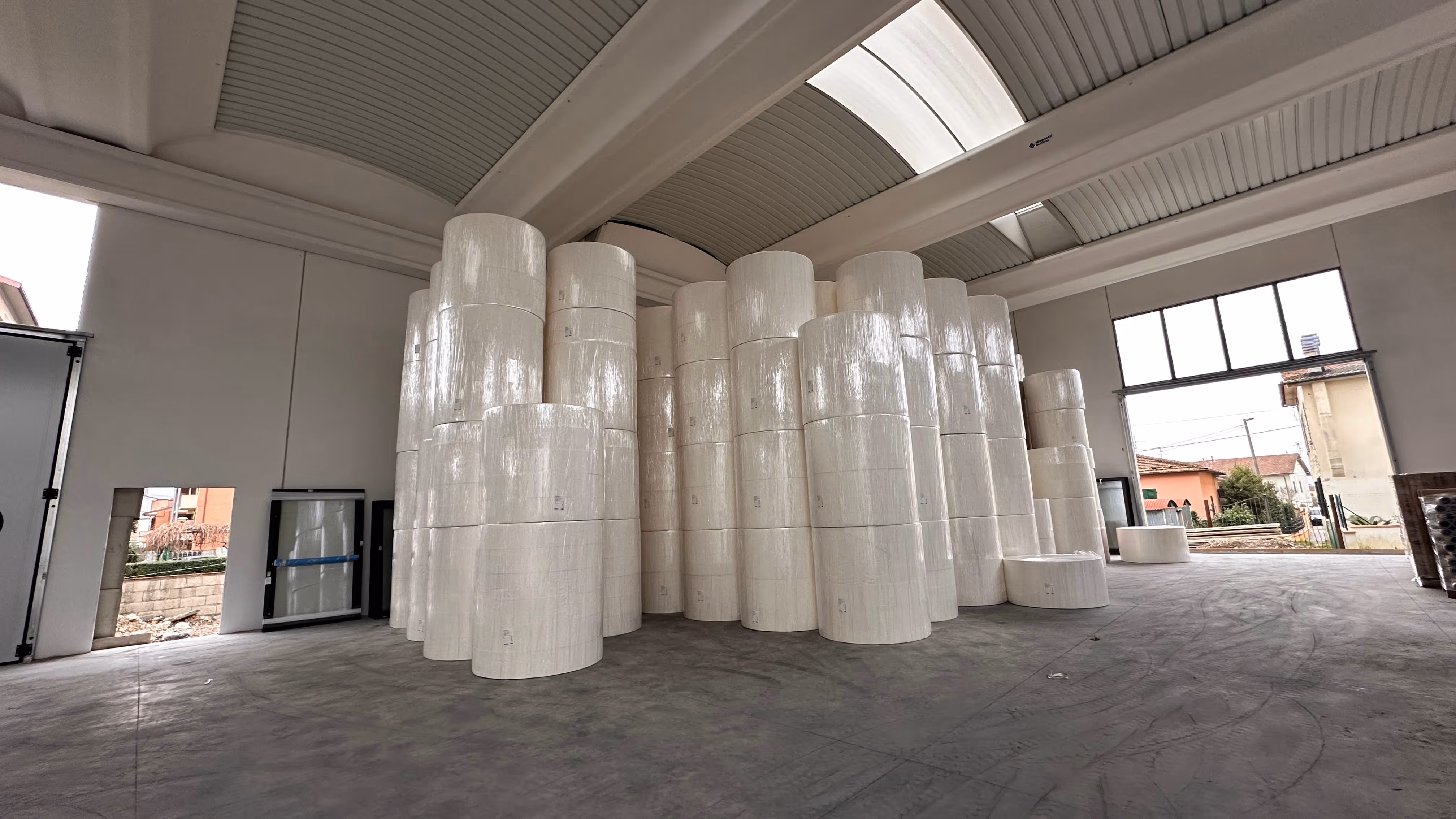 Grande magazzino con numerosi grandi rotoli di carta bianca impilati al centro su pavimento in cemento.