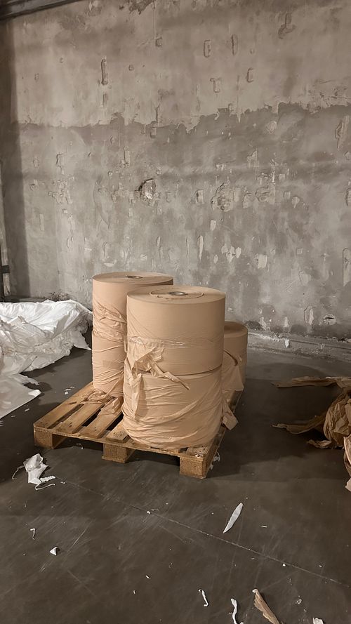 Tre grandi rotoli di carta avvolti con carta marrone, posizionati su un bancale di legno in un magazzino con pareti di cemento grezzo.