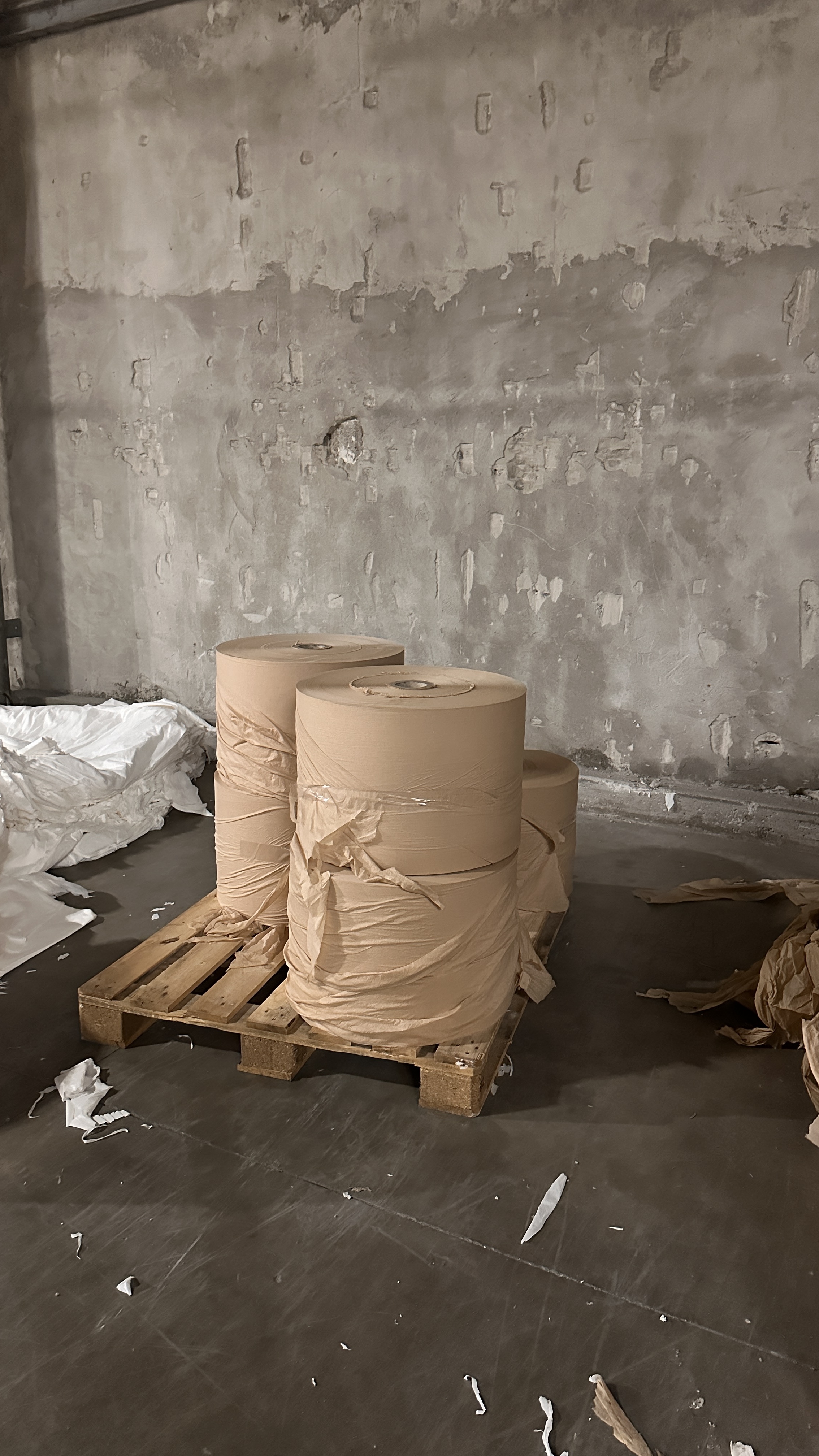 Tre grandi rotoli di carta avvolti con carta marrone, posizionati su un bancale di legno in un magazzino con pareti di cemento grezzo.