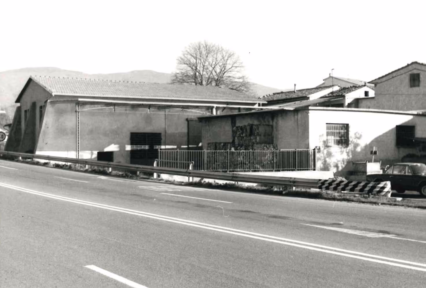 Edifici industriali con recinzione metallica lungo una strada asfaltata con linea di mezzeria doppia, auto parcheggiata sul lato destro.