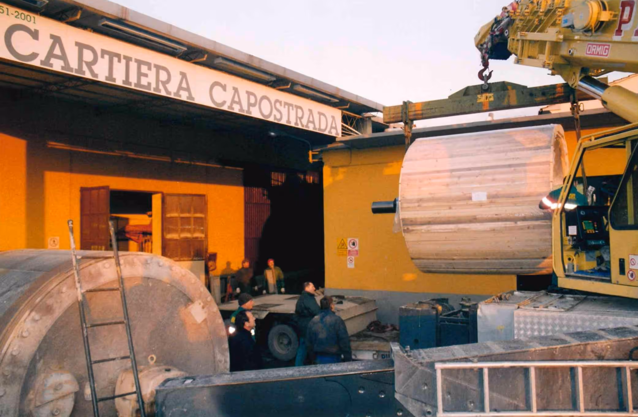 Operai in una cartiera mentre una gru solleva un grande rotolo di carta all'esterno di un edificio giallo con insegna Cartiera Capostrada.