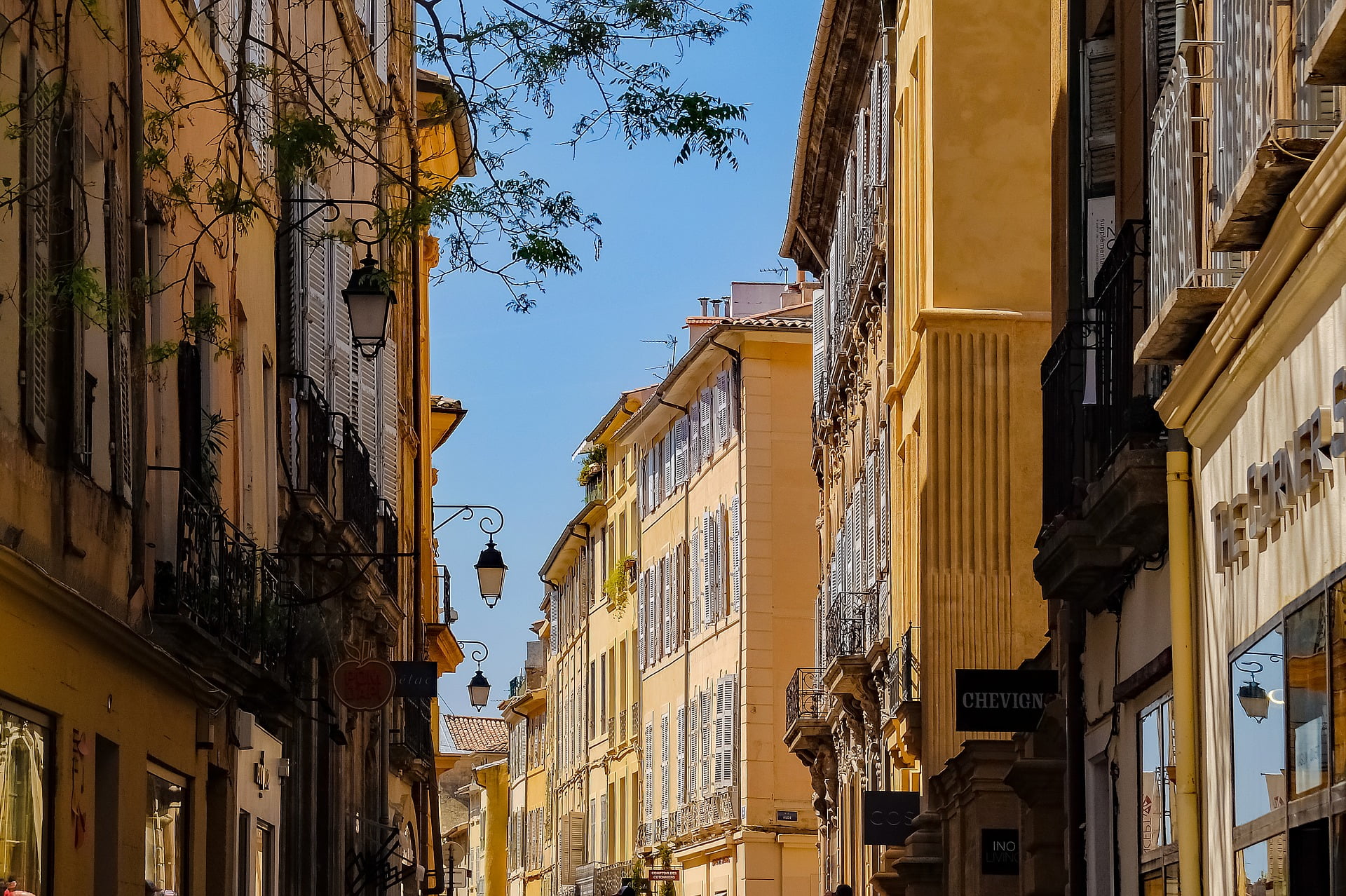 Narrow European street lined with tall, historic buildings featuring shuttered windows under a clear blue sky.