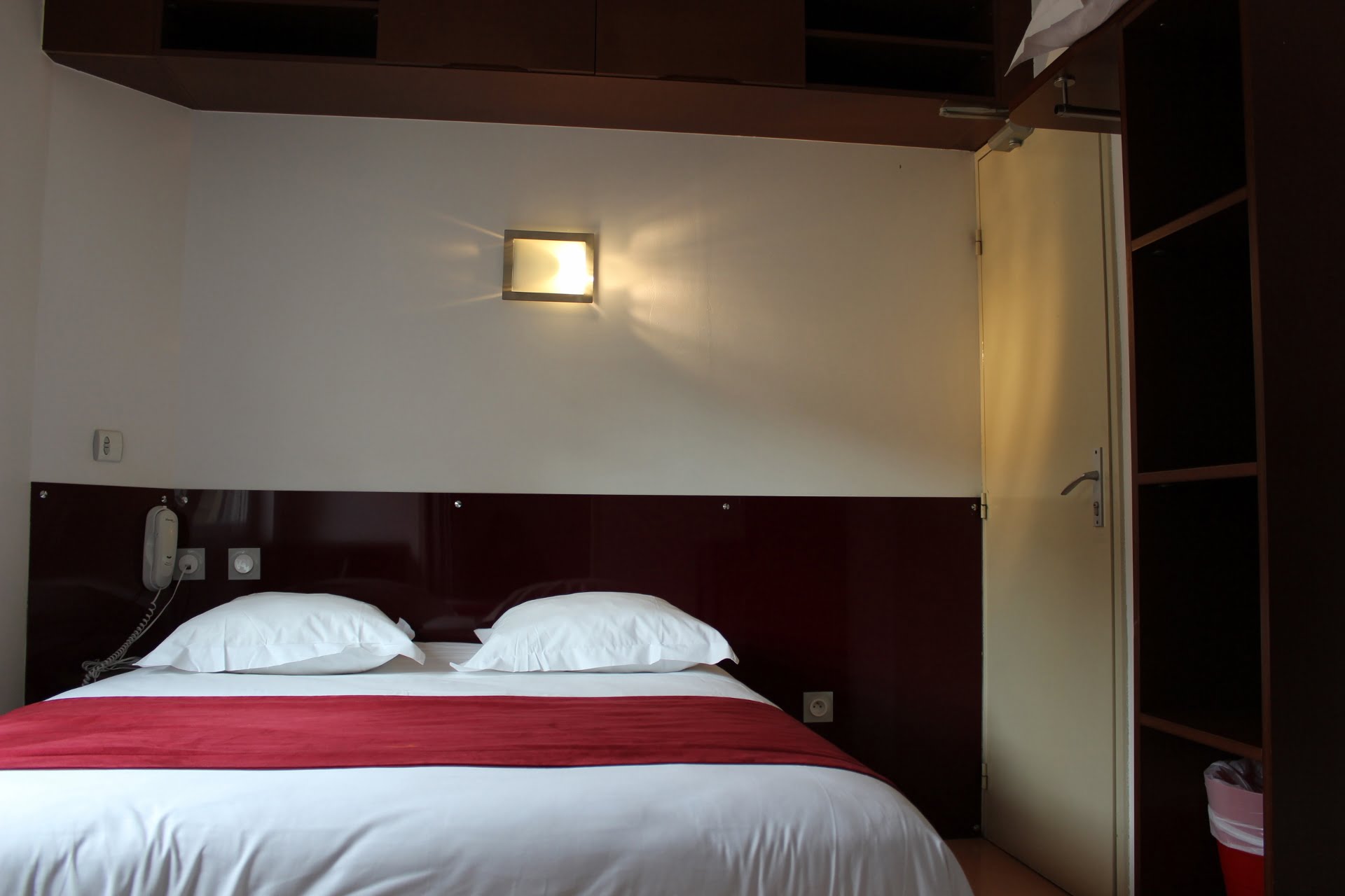 Minimalist hotel room with a double bed featuring white linens and a red runner, a wall-mounted light, closed door, and built-in shelving.