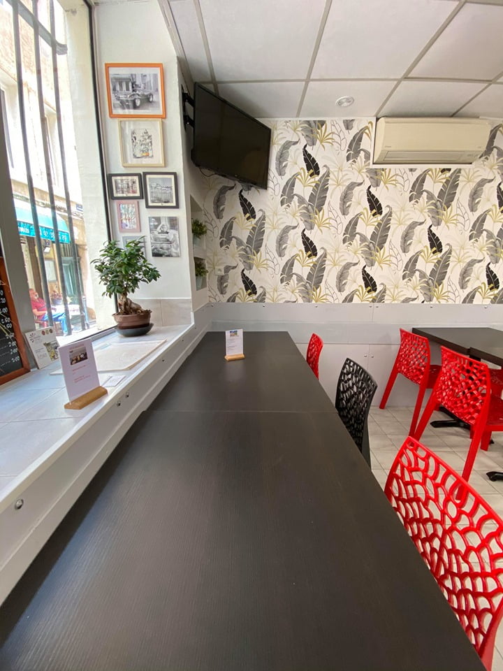 Modern cafe interior with black tables, red and black lattice-back chairs, wall-mounted TV, leafy wallpaper, and window seating with potted plant.