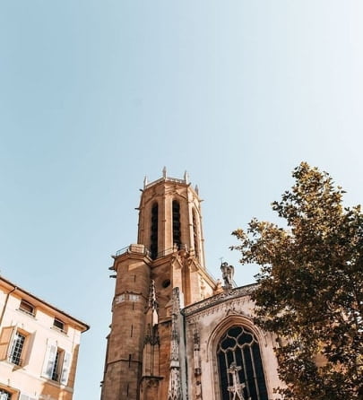 Low angle view of a historic stone church tower with gothic windows and a tree in the foreground under a clear blue sky.