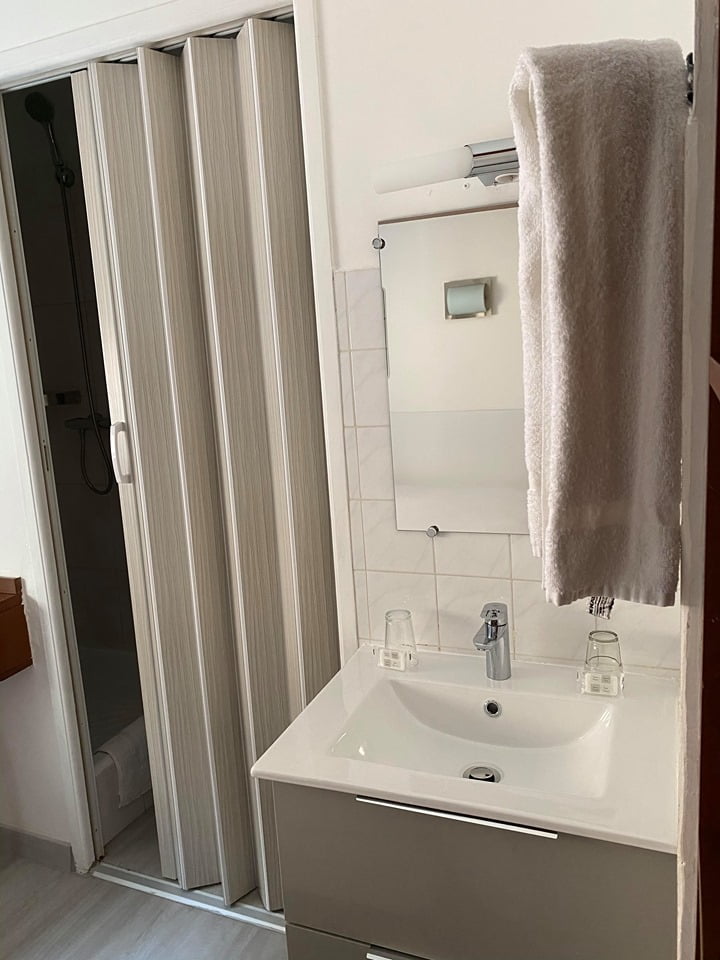 Compact bathroom sink with two glasses, a faucet, wall-mounted mirror, white towel hanging, and a partially open folding door revealing a shower area.