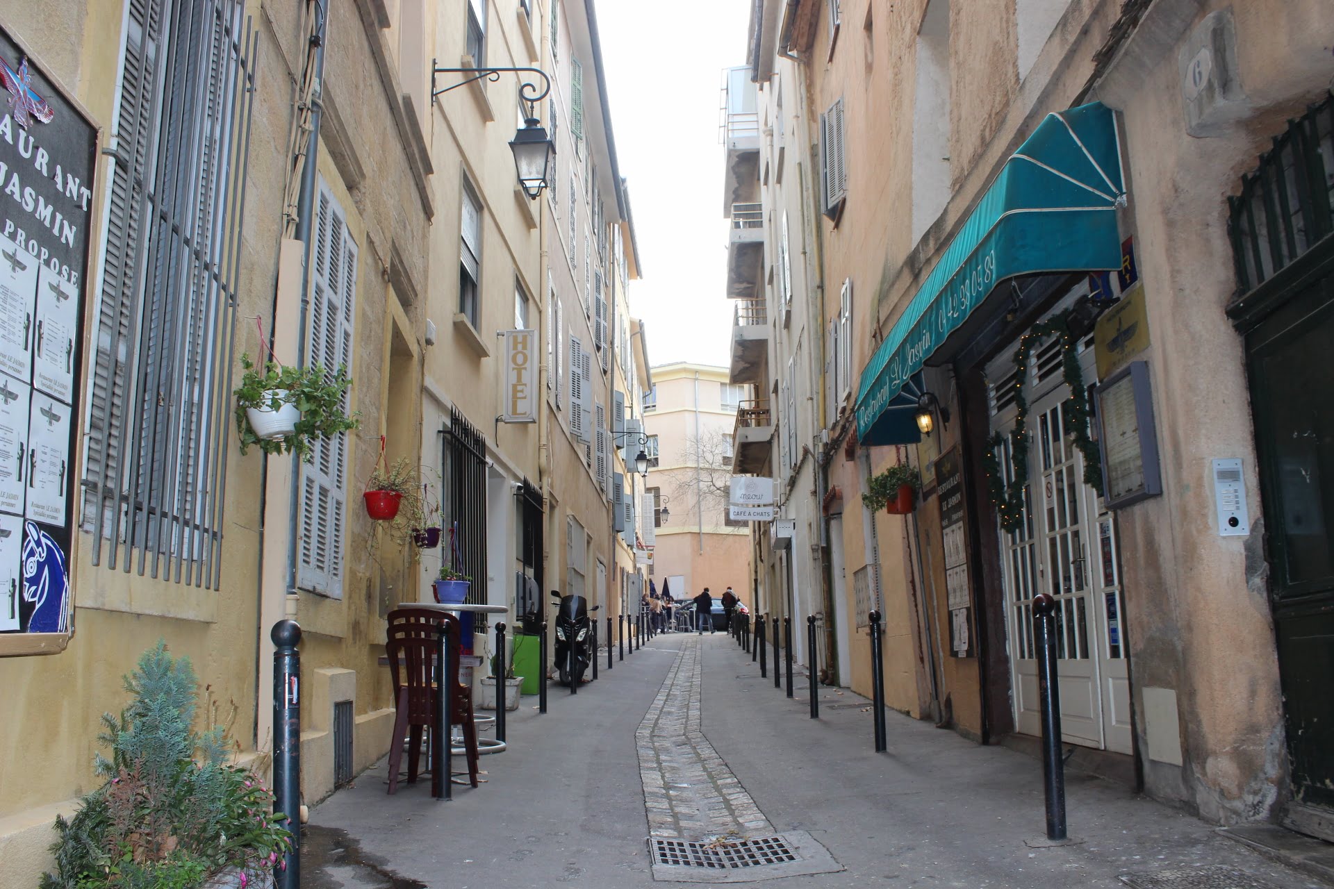 Narrow European street lined with cafes and shops, outdoor tables, hanging plants, and a hotel sign.