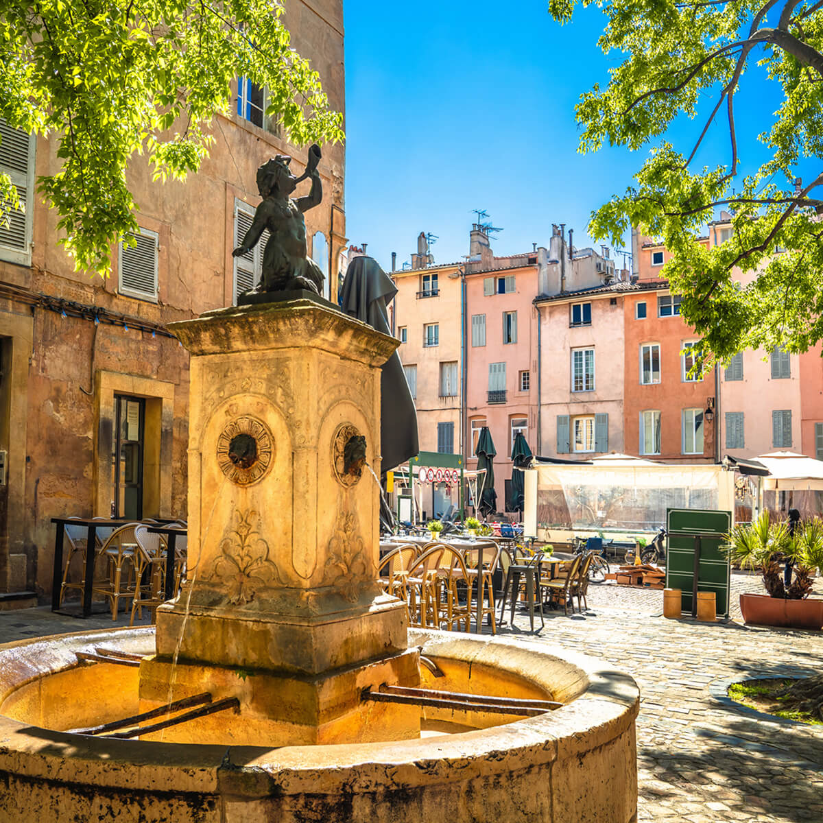 Sunlit stone fountain with a bronze statue of a figure blowing a horn in a cobblestone square with outdoor cafe seating and pastel buildings.