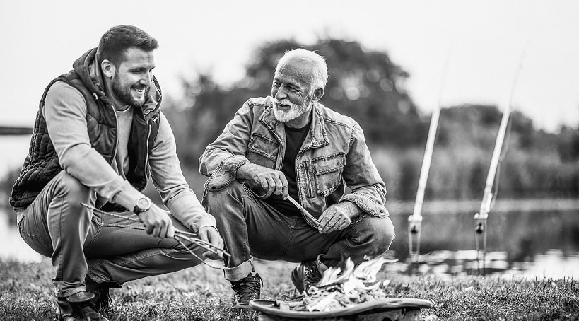 Retired man and his son sitting around a campfire by a lake.
