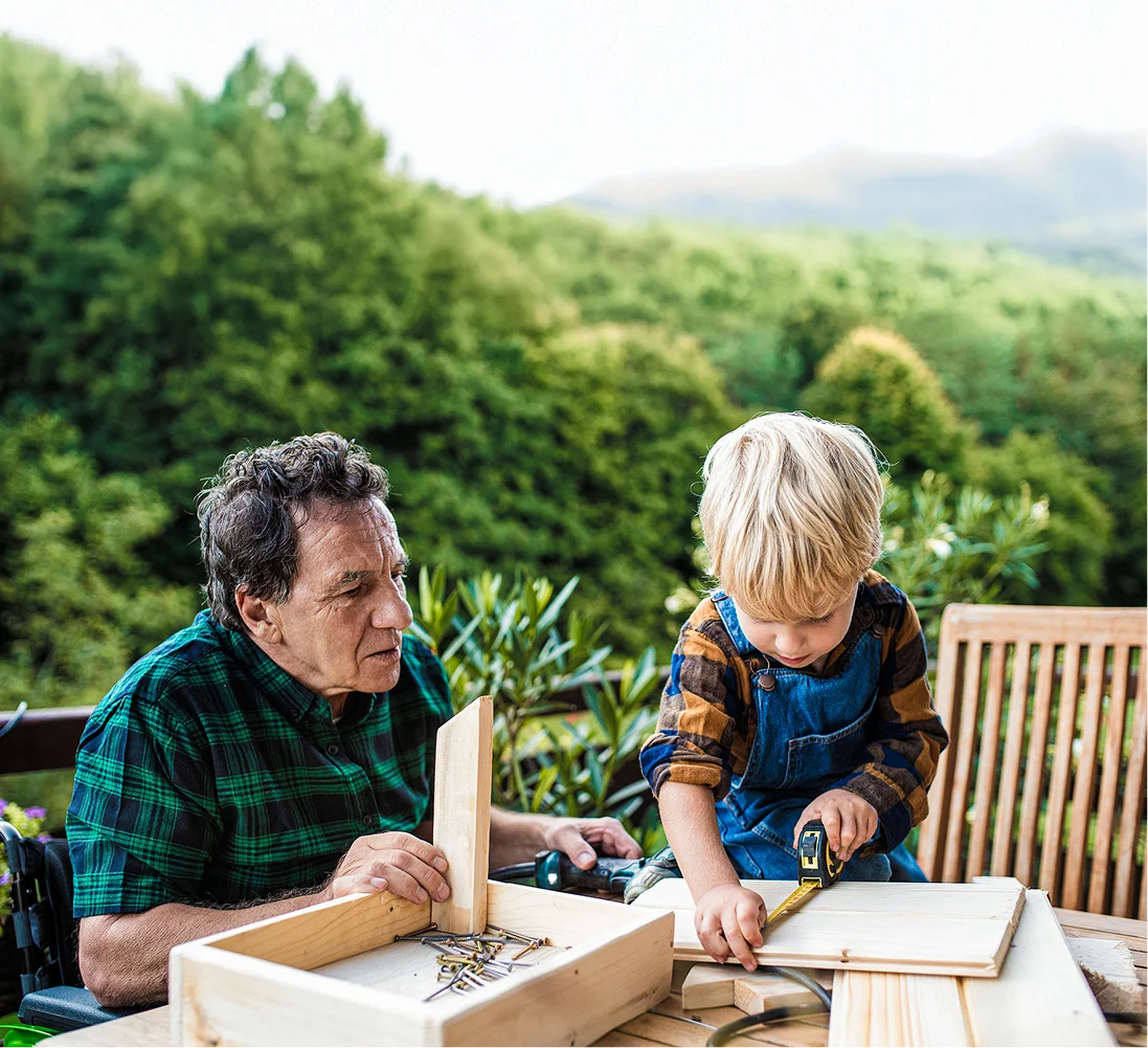 Retired man watches grandson using a tape measure on wood for a woodworking project outdoors.