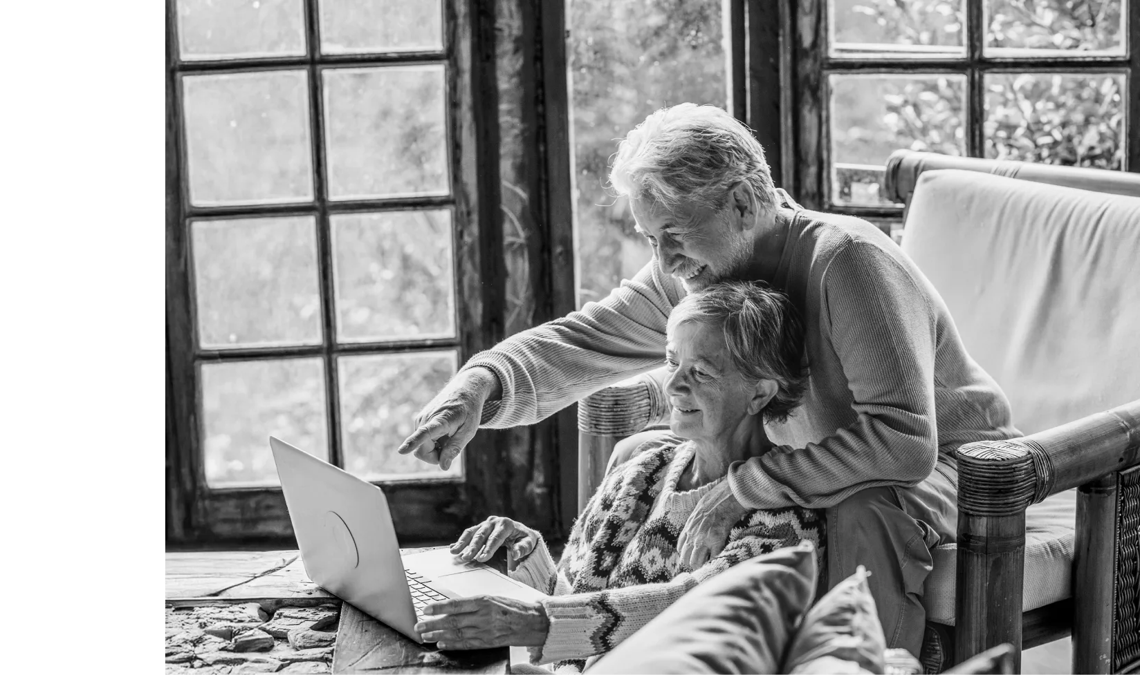 Smiling retired couple using laptop together in a cozy room with large windows.