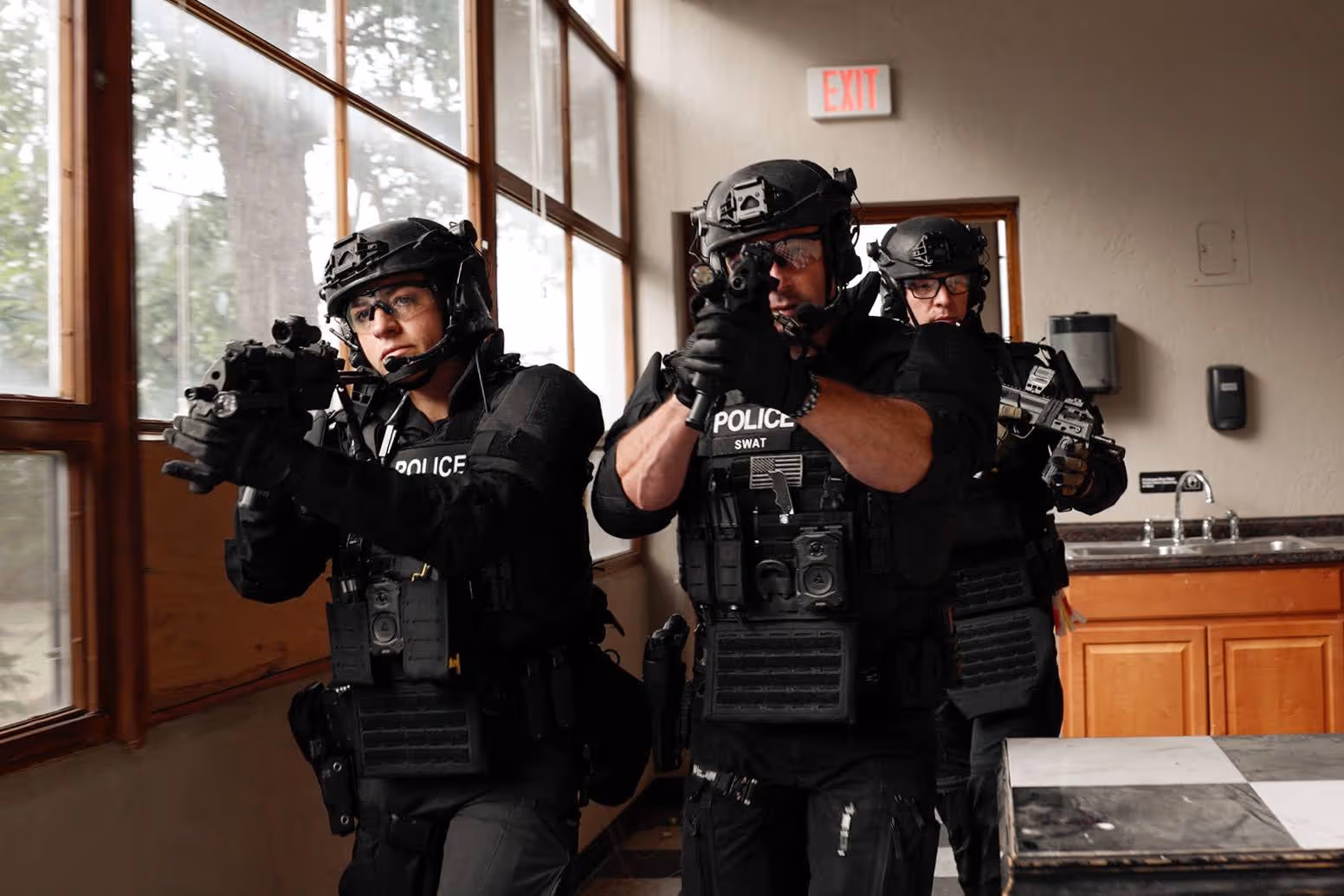 Three police SWAT officers in tactical gear aiming rifles inside a building near windows and an exit sign.