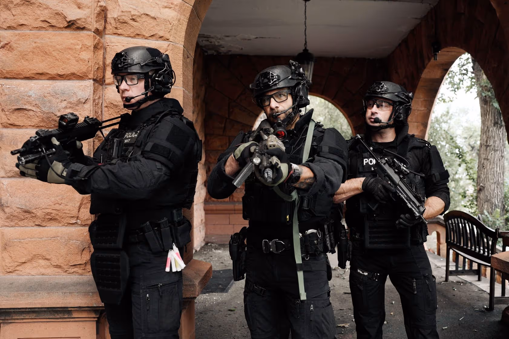 Three police officers in tactical gear with rifles positioned under a stone archway.