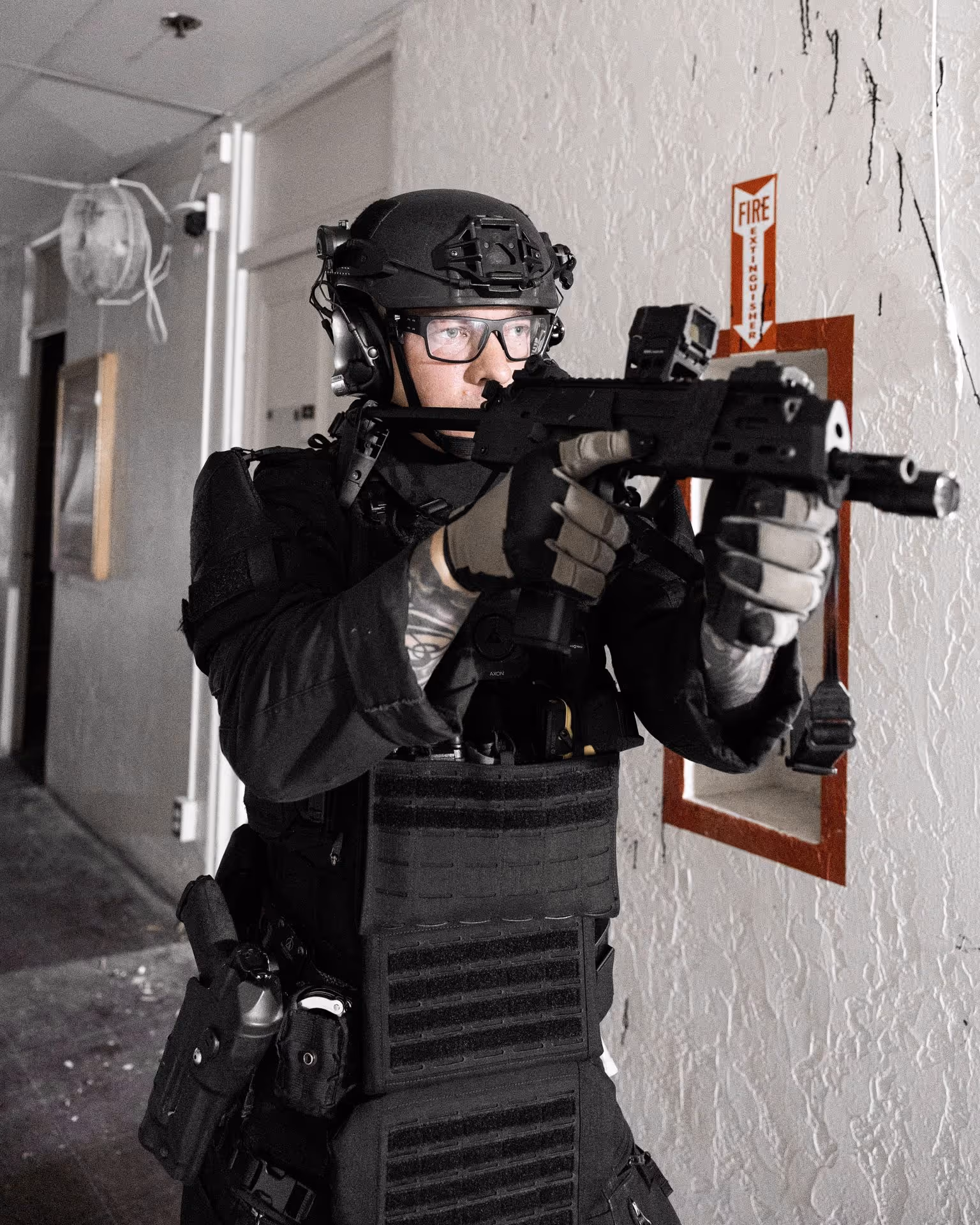 Tactical officer in black gear aiming a rifle indoors near a fire extinguisher sign on a white wall.