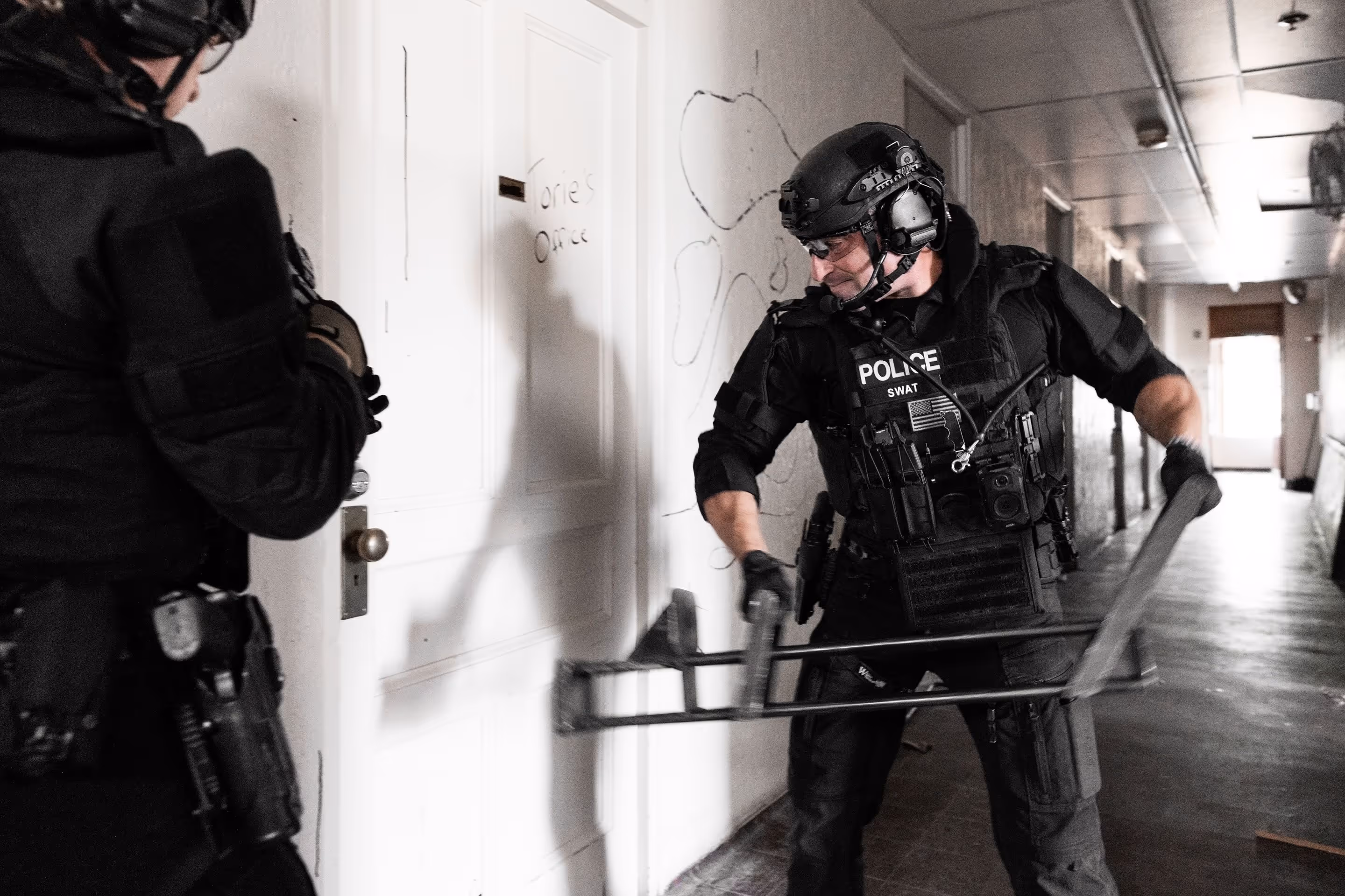 SWAT officers in tactical gear prepare to breach a white door in a dim hallway.