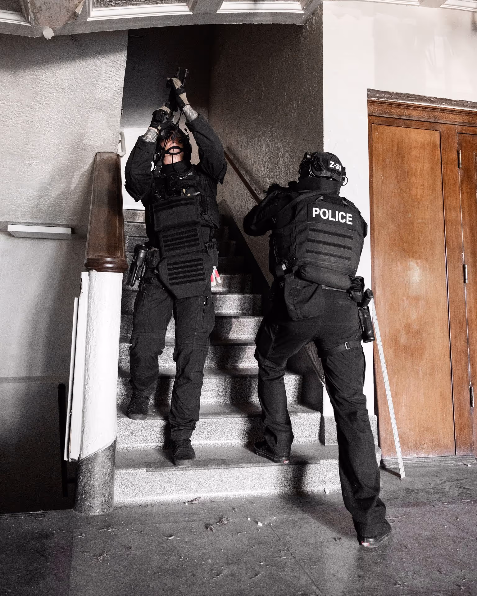 Two police officers in tactical gear ascending a staircase inside a building, one aiming a rifle upwards and the other covering the stairway.