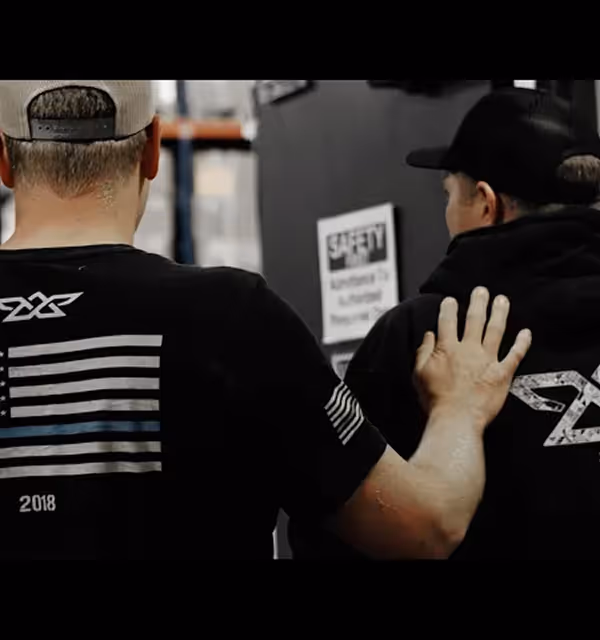 Man with hand on another's shoulder, both wearing black shirts with logos in an indoor setting.