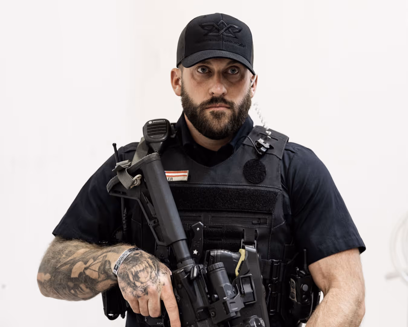 Tattooed man in tactical gear holding a rifle, wearing a black cap and radio equipment.
