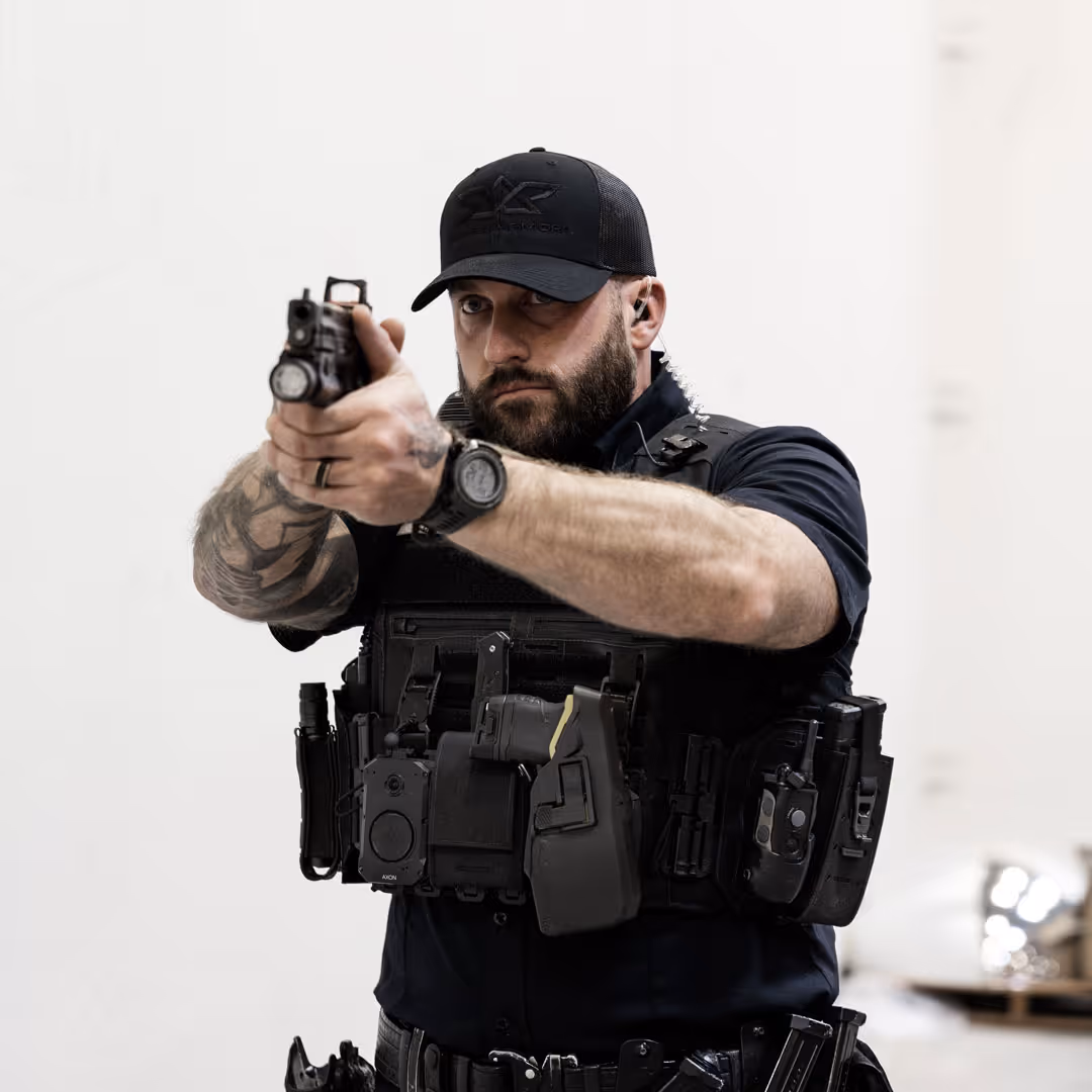 Tactical police officer aiming handgun forward, wearing black gear and cap, with tattoos on left arm.