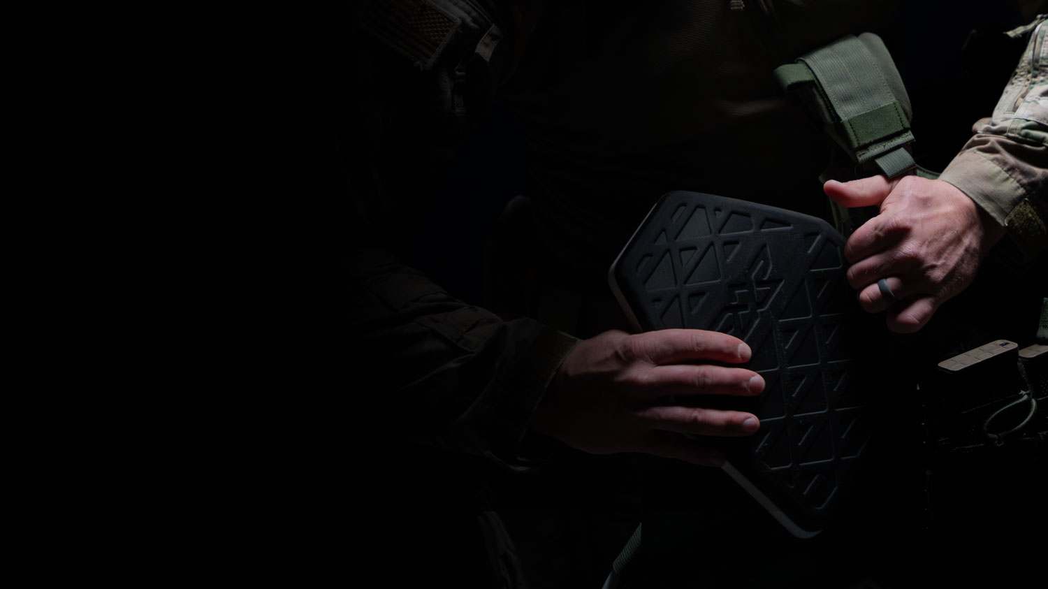 Person in tactical gear holding a black protective armor plate with geometric patterns.
