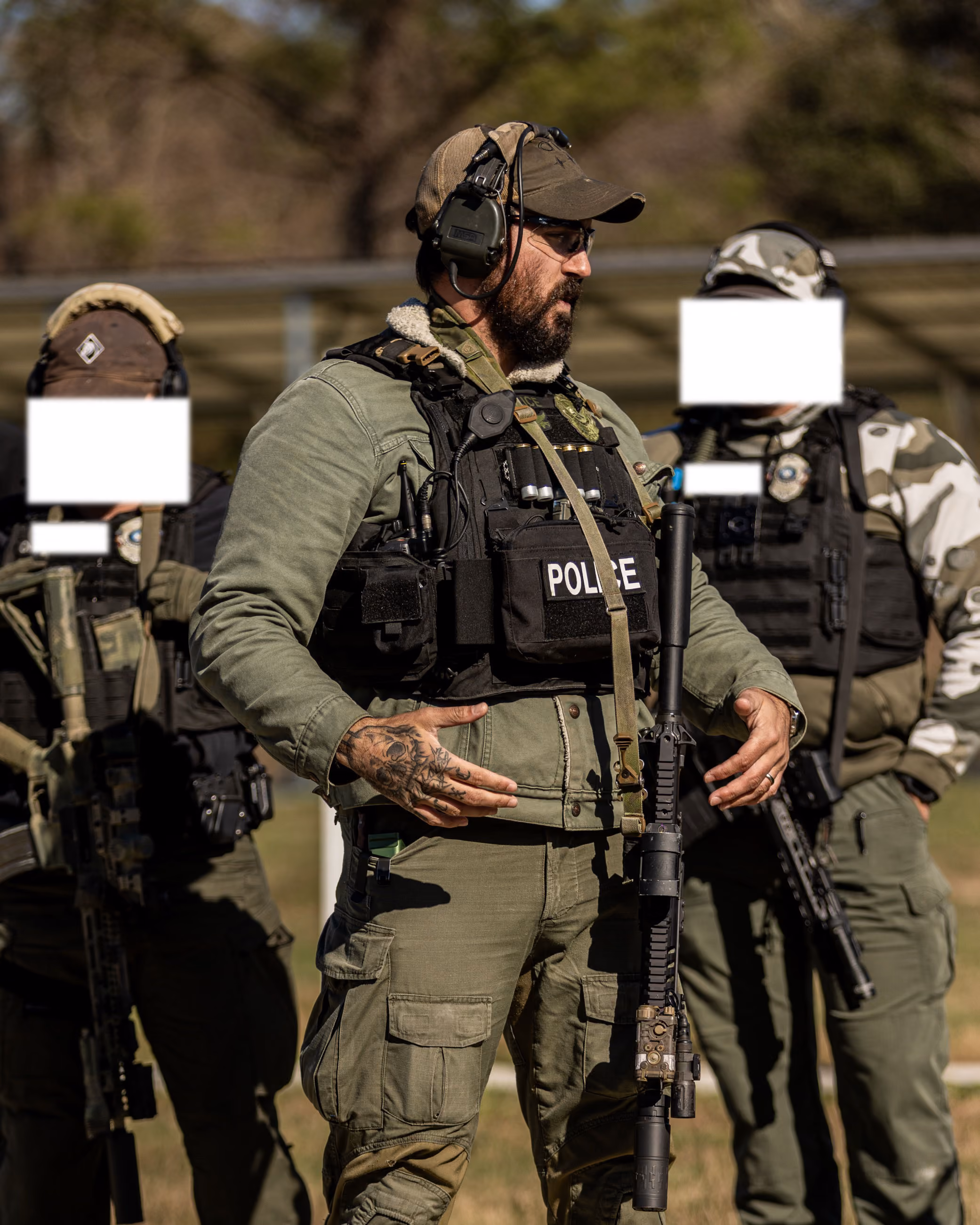 Tattooed police officer in tactical gear speaks with two armed officers in outdoor training setting.